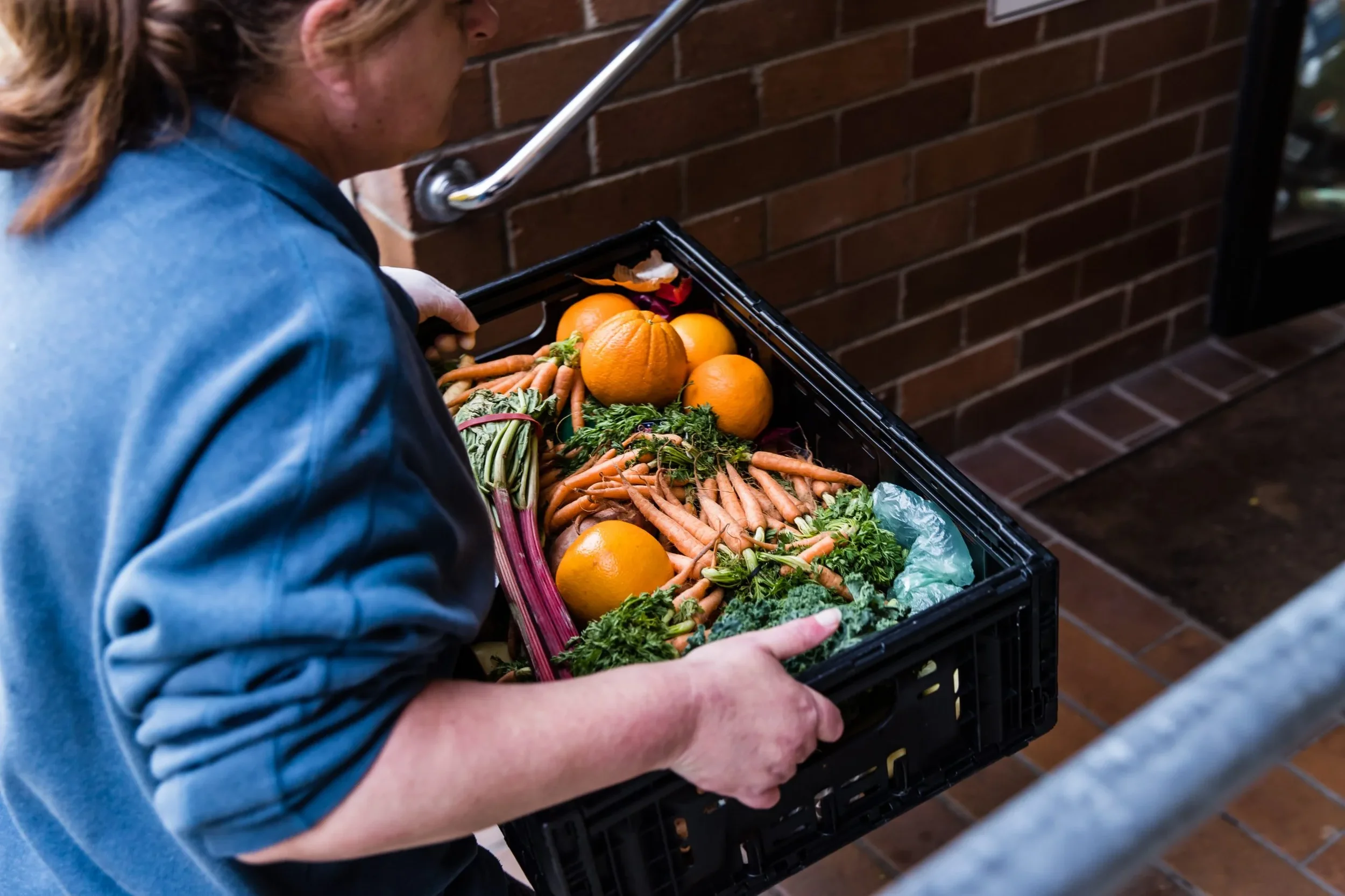 Person wearing a blue jacket holding a black crate filled with oranges, carrots, and leafy greens outside near a brick wall.