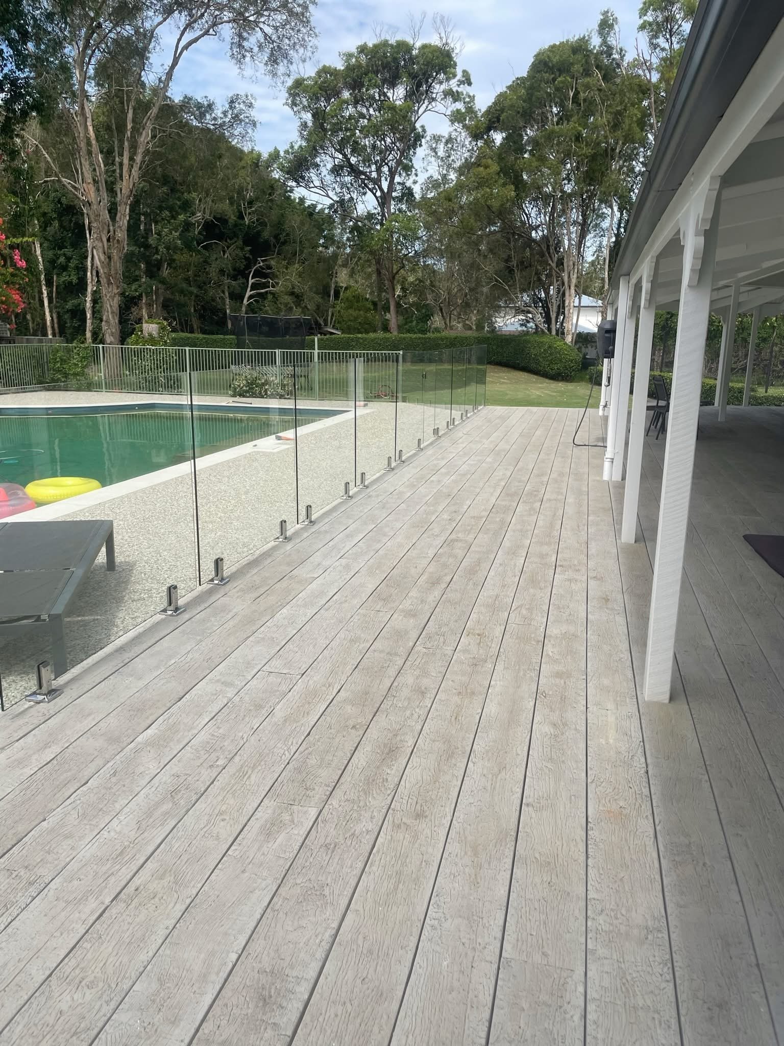 Outdoor backyard with a swimming pool, glass safety fencing, a wooden deck, and trees in the background.