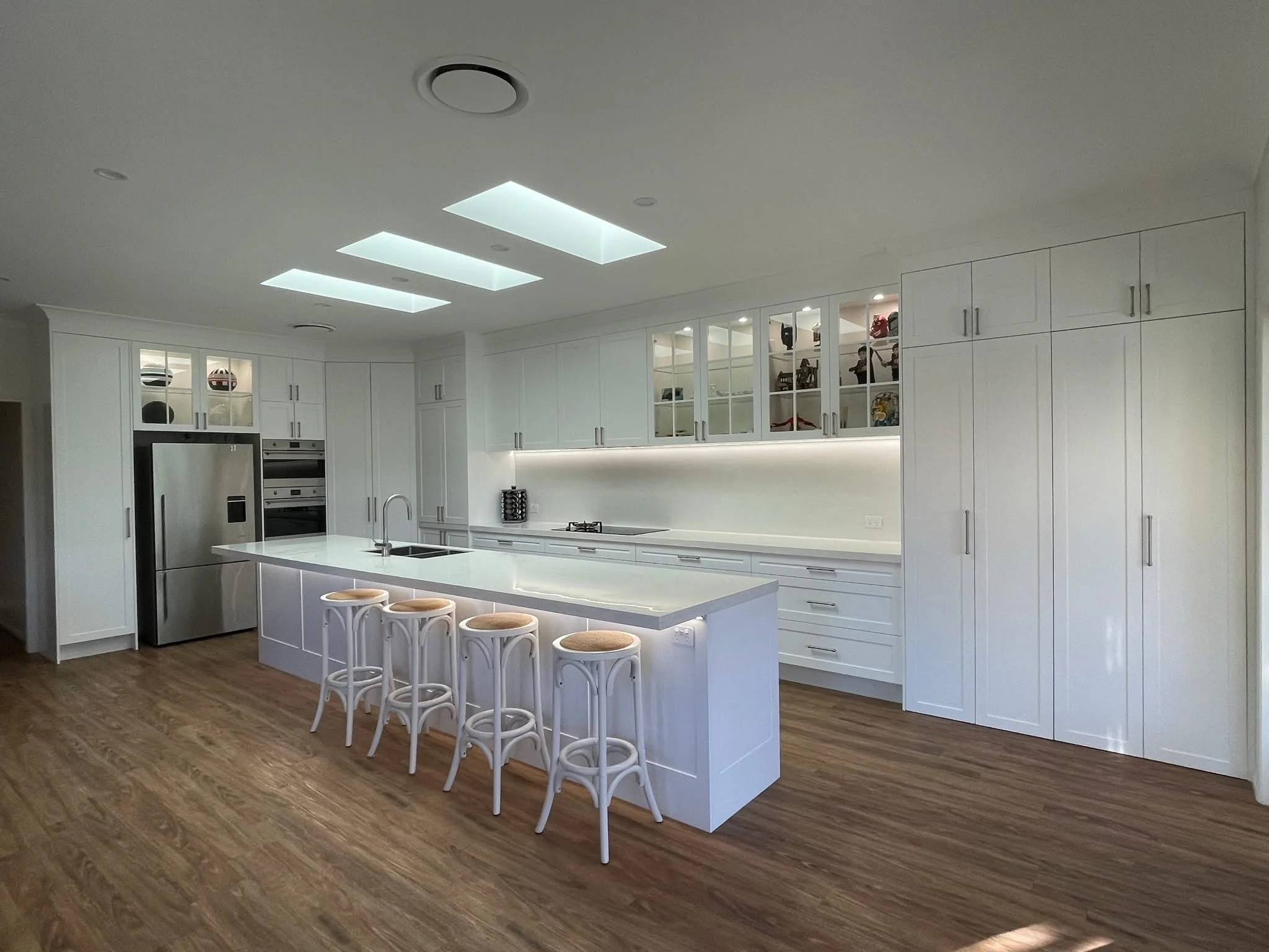 Modern kitchen with white cabinets, stainless steel refrigerator, central island with four stools, and wood flooring, under a ceiling with rectangular skylights.