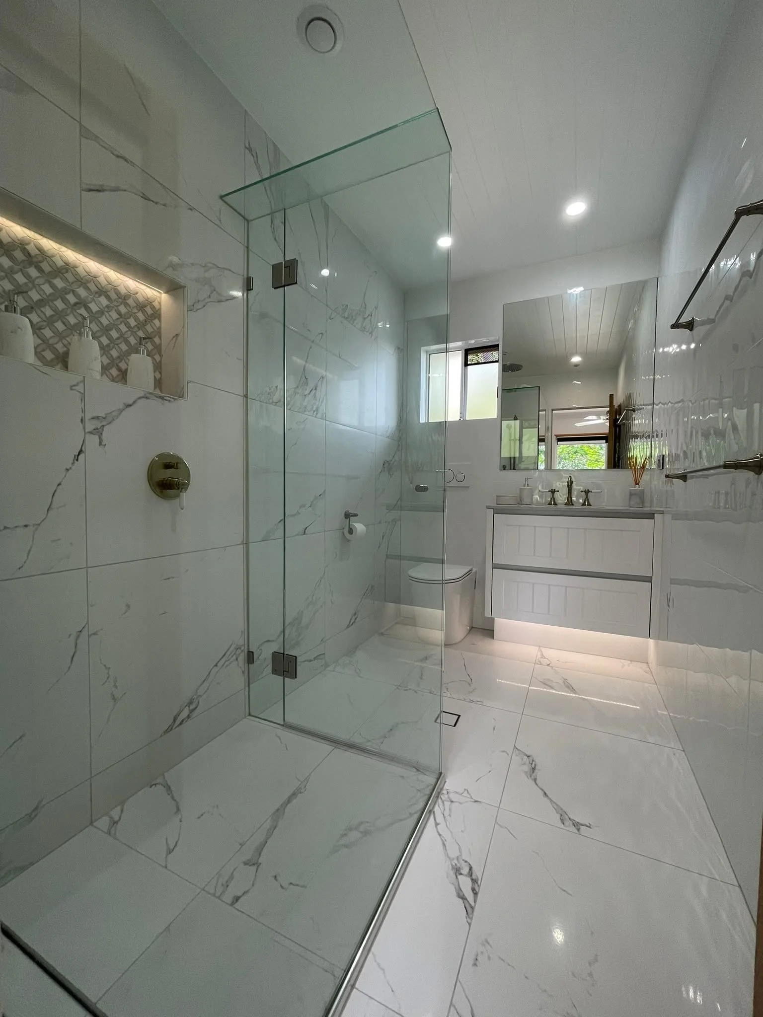 Modern bathroom with marble tiles, glass shower enclosure, vanity with sink, and large mirror, illuminated by ceiling lights and natural light from window.