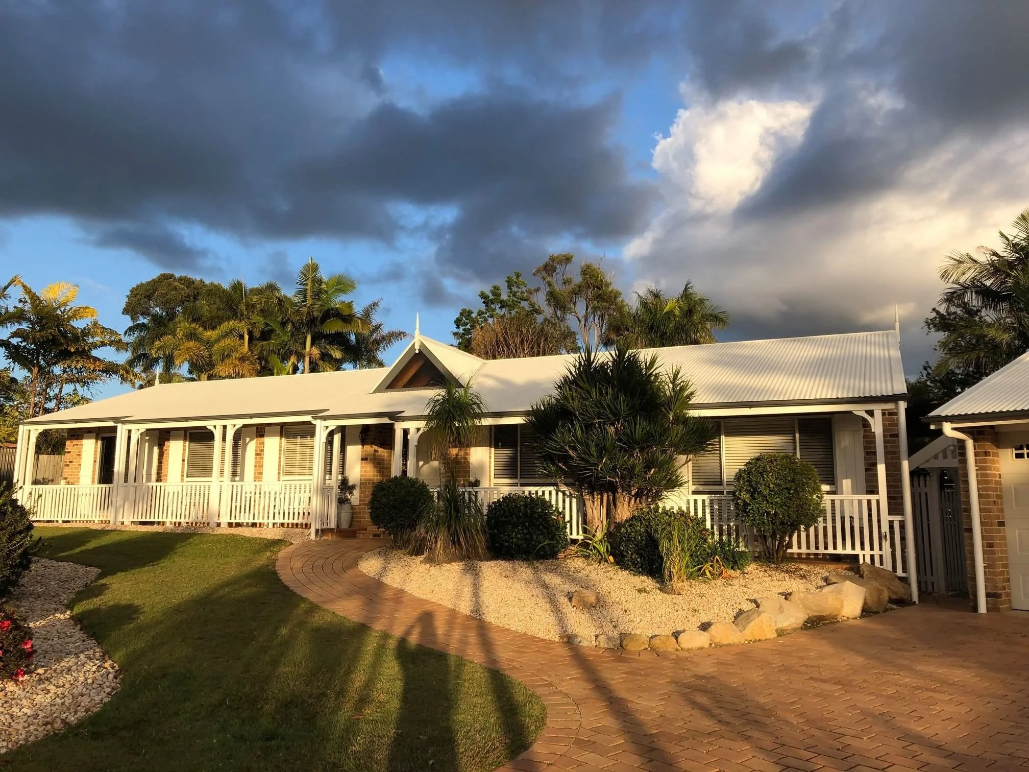 A house with a white roof and brick pillars, surrounded by a landscaped yard with green grass, trees, bushes, and a curved brick pathway, under a sky with dark and light clouds.