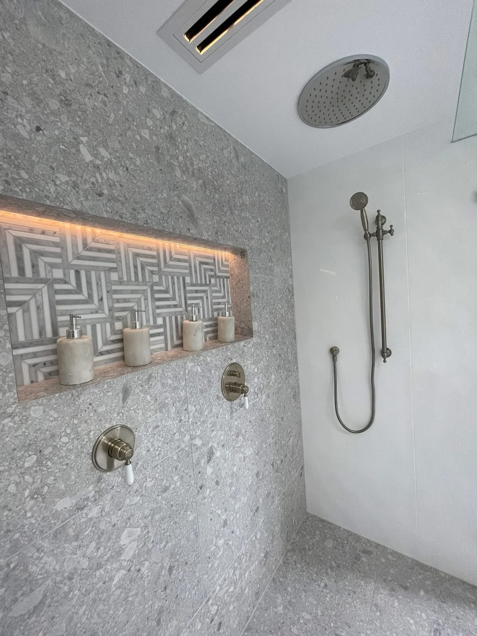 A modern shower area with a gray speckled wall, a decorative niche with black, white, and gray geometric tiles, four soap dispensers, a ceiling-mounted rainfall showerhead, a handheld shower on a rail, and an overhead ventilation grille.