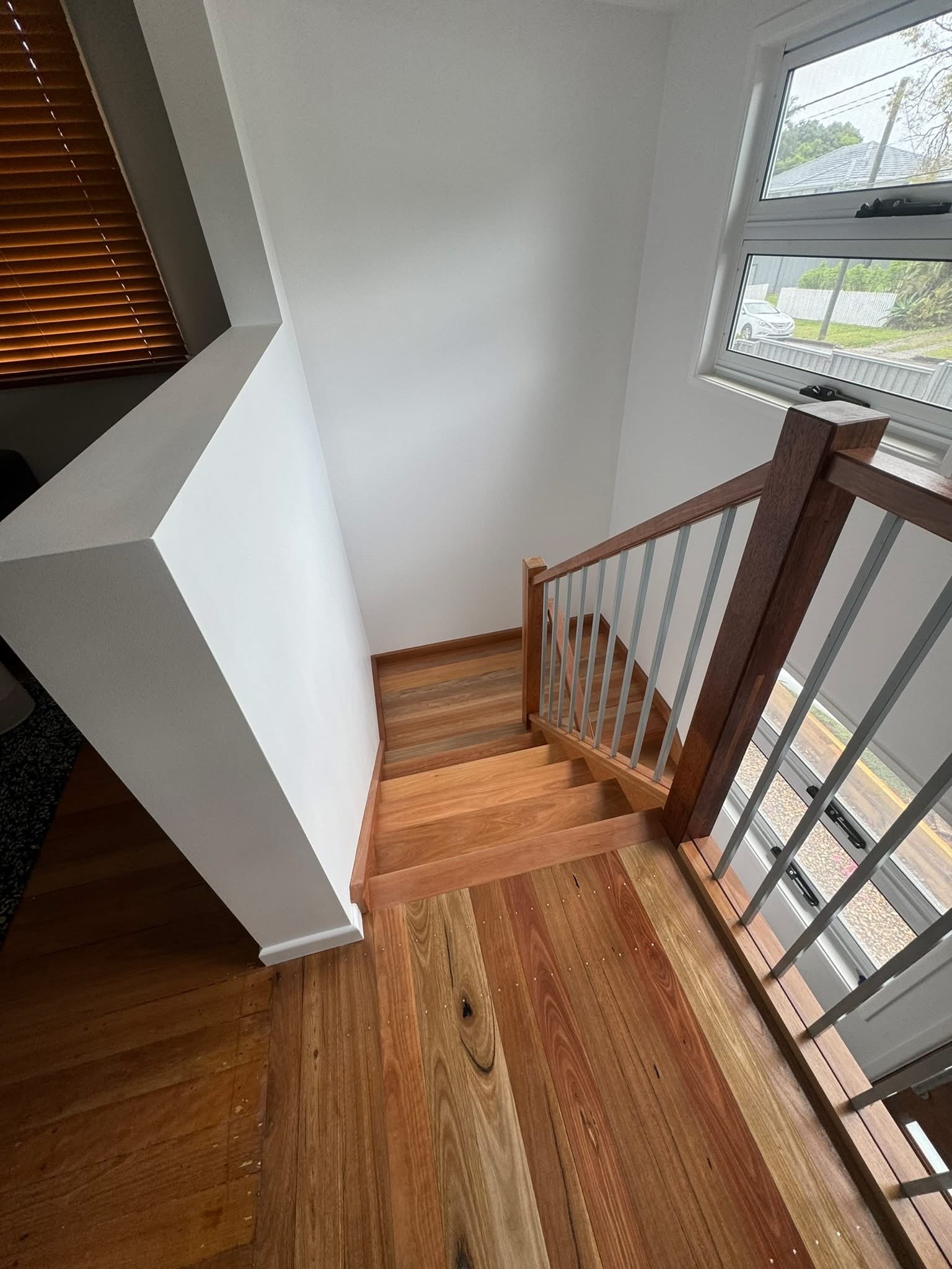 Interior view of a staircase with wooden steps and handrails, white walls, and a window showing a view outside with a car and greenery.