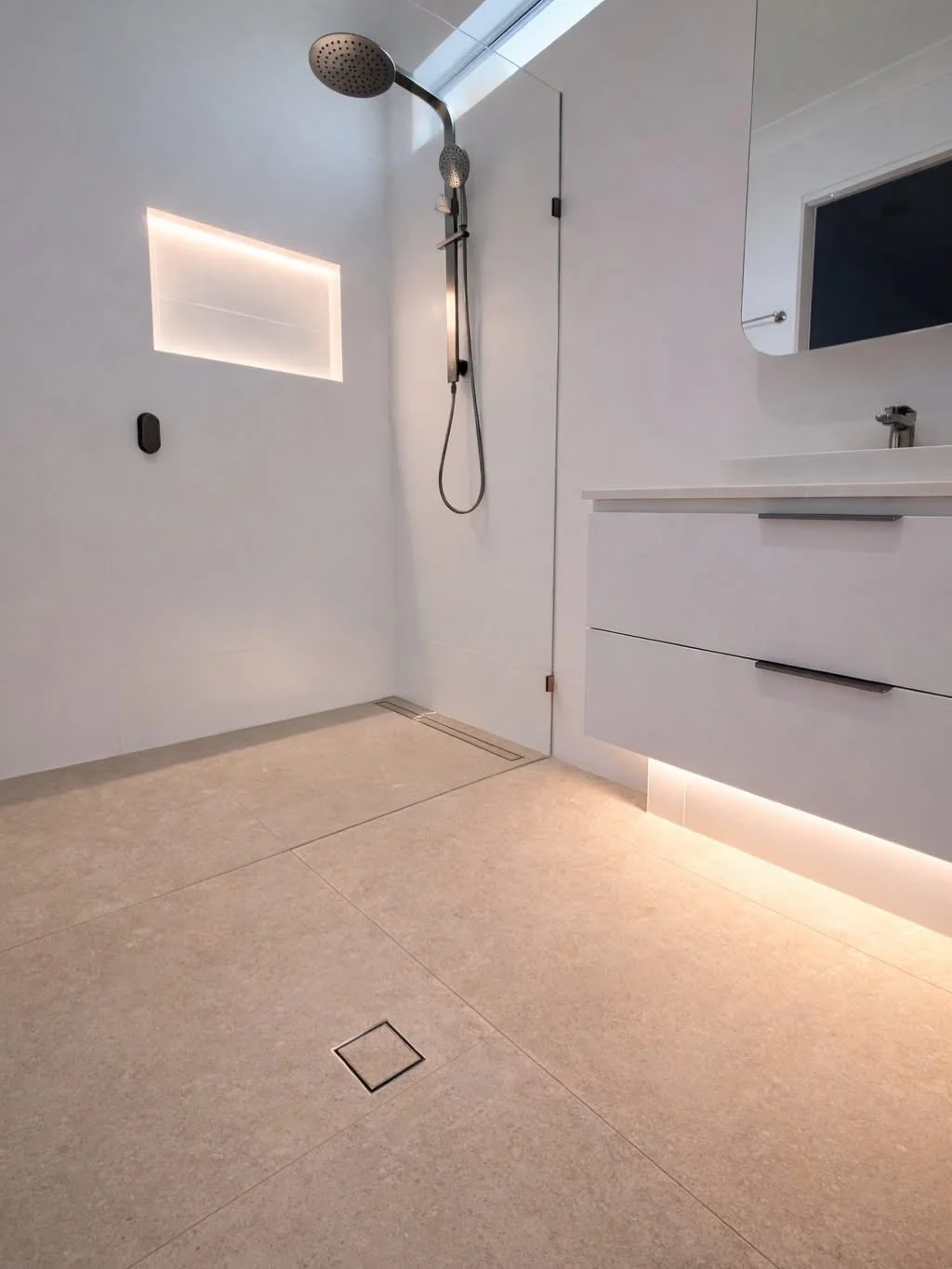 Modern bathroom shower area with white walls, beige tiled floor, and a wall-mounted shower with a silver showerhead and hand-held spray. There's a built-in niche with backlighting and a white vanity with drawer storage and a faucet. A mirror is above the sink, and a rectangular drain is in the floor.