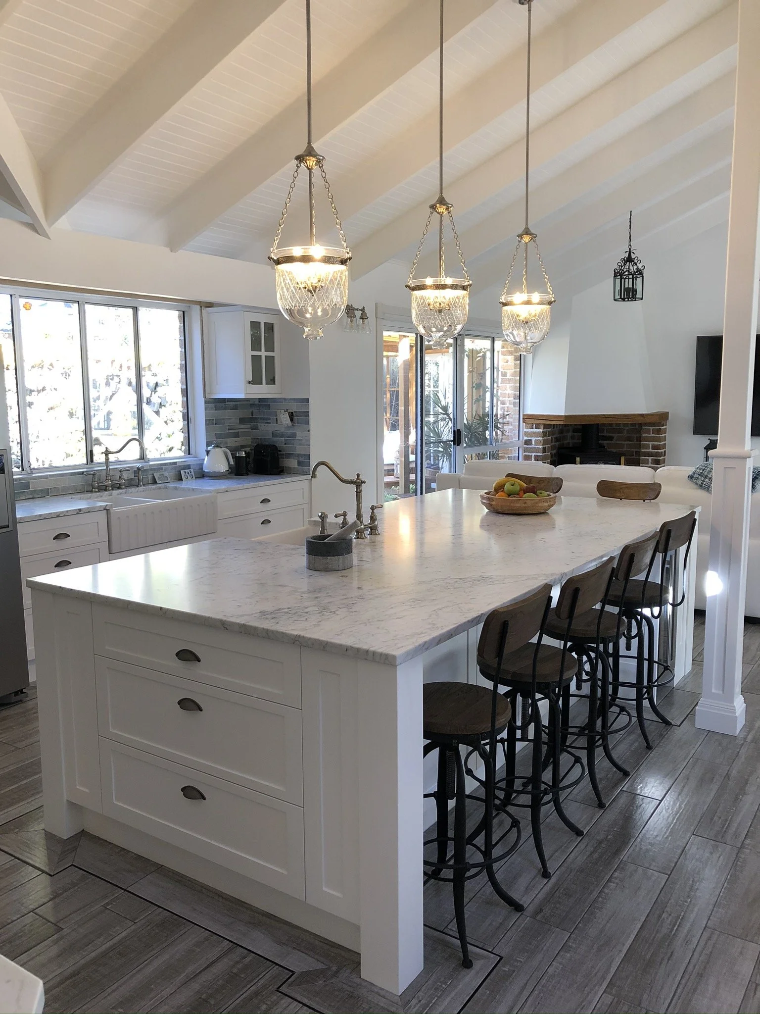 A modern kitchen with a large white island with drawers and a marble countertop, black bar stools, pendant lights hanging from a vaulted ceiling, a window over the sink, and a cozy brick fireplace in the background.