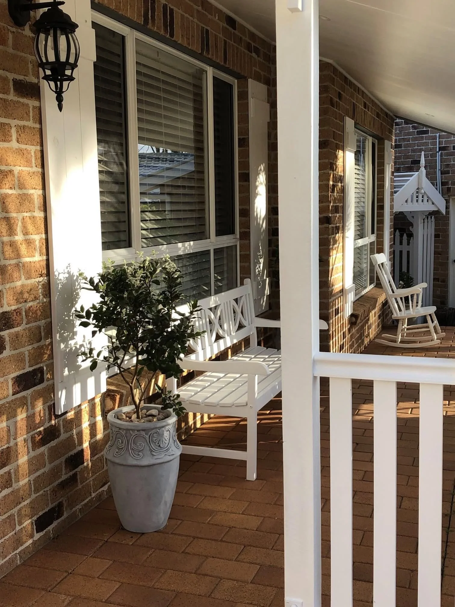 A porch with a brick wall, white railings, a white bench, two white rocking chairs, and a potted plant in a decorative white pot. A black porch lantern is mounted on the wall.