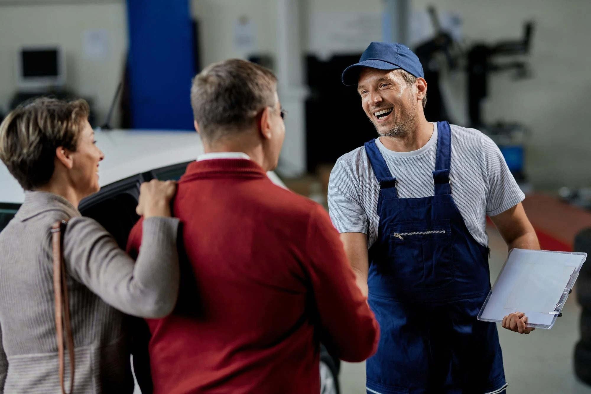 Three people engaged in conversation indoors, with a worker in a blue cap and overalls smiling while talking to two women.
