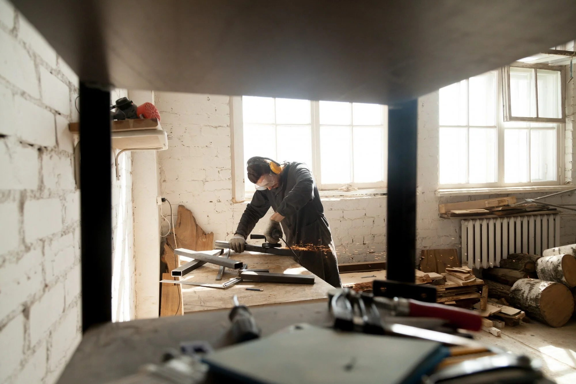 A person wearing safety goggles, a face mask, and gloves is welding metal pieces on a workbench in a workshop with brick walls and large windows.