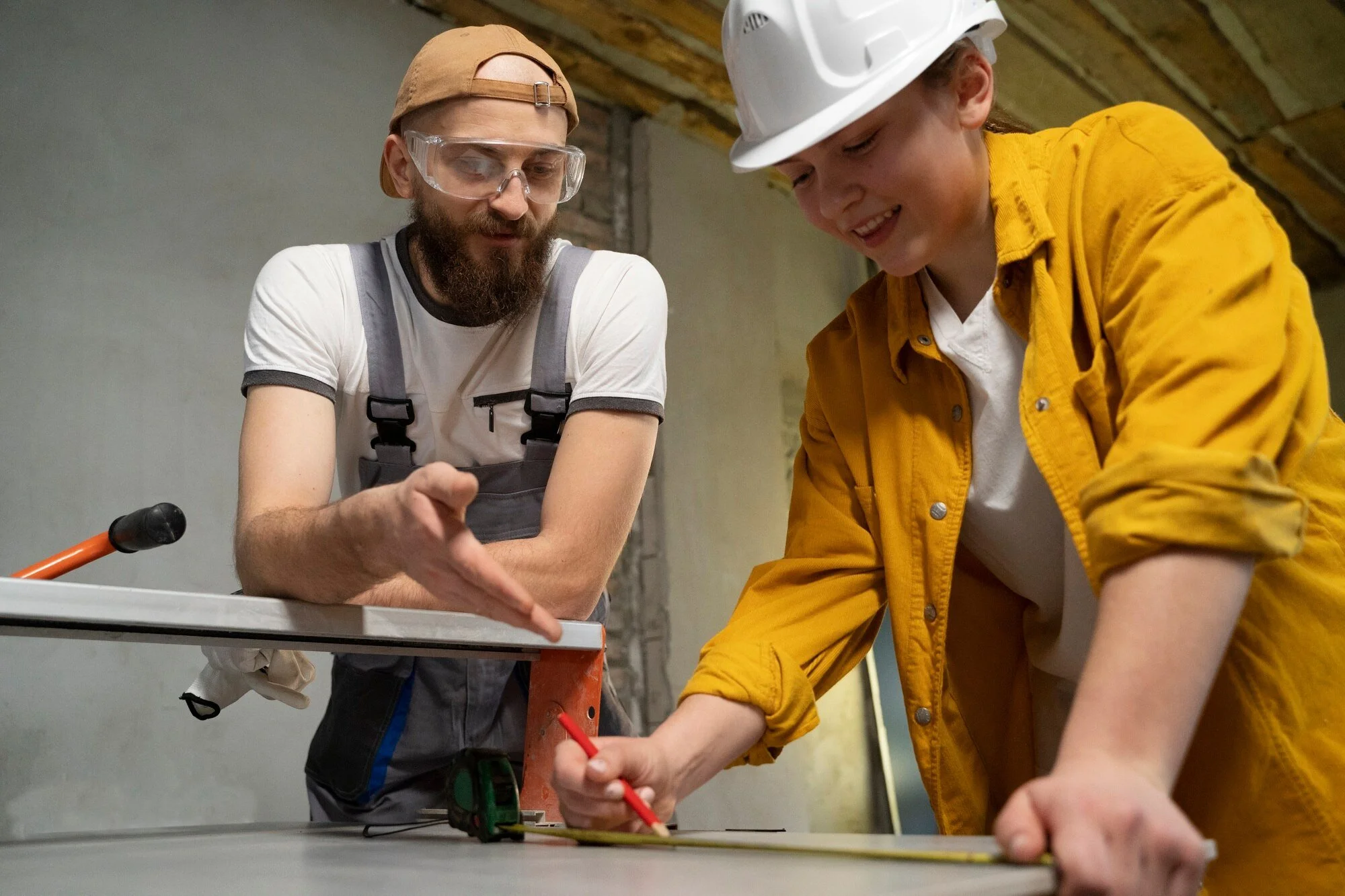 Two people, a man with a beard wearing transparent safety goggles and a beige cap, and a woman wearing a white hard hat and a yellow jacket, are working together on a construction project. They are focused and smiling, using a red tool on a piece of wood in an indoor setting with an unfinished ceiling.