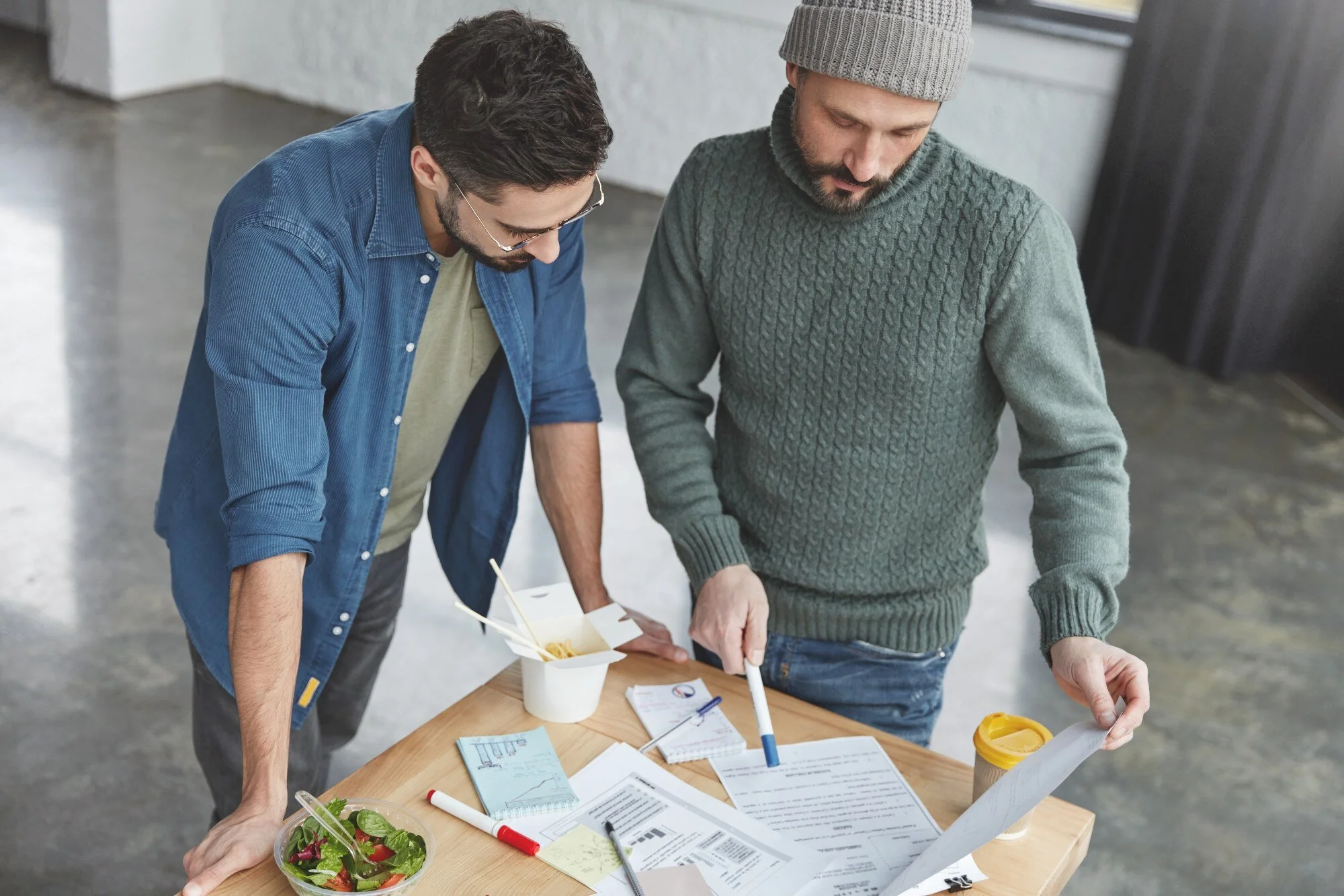 Two men standing at a table reviewing papers, with one holding a document and a container of salad, on a wooden table with takeout food and office supplies, in an industrial-style room.