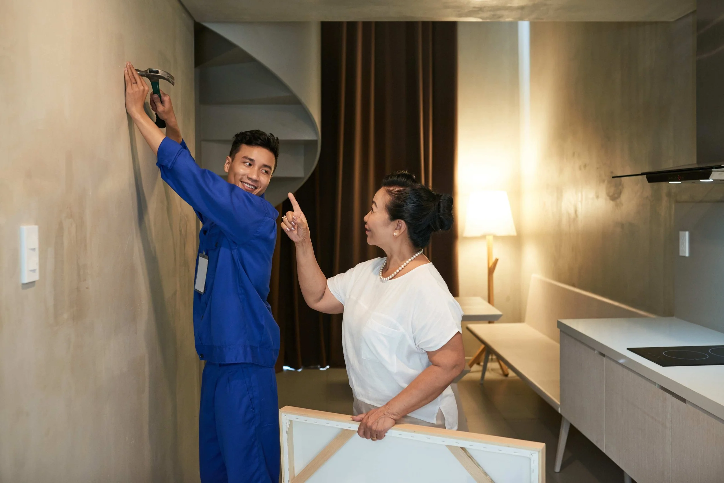 A young man in blue scrubs hammering a nail into a beige wall while an older woman in white blouse watches, pointing at him, in a room with a lamp, brown curtains, and a kitchen counter.