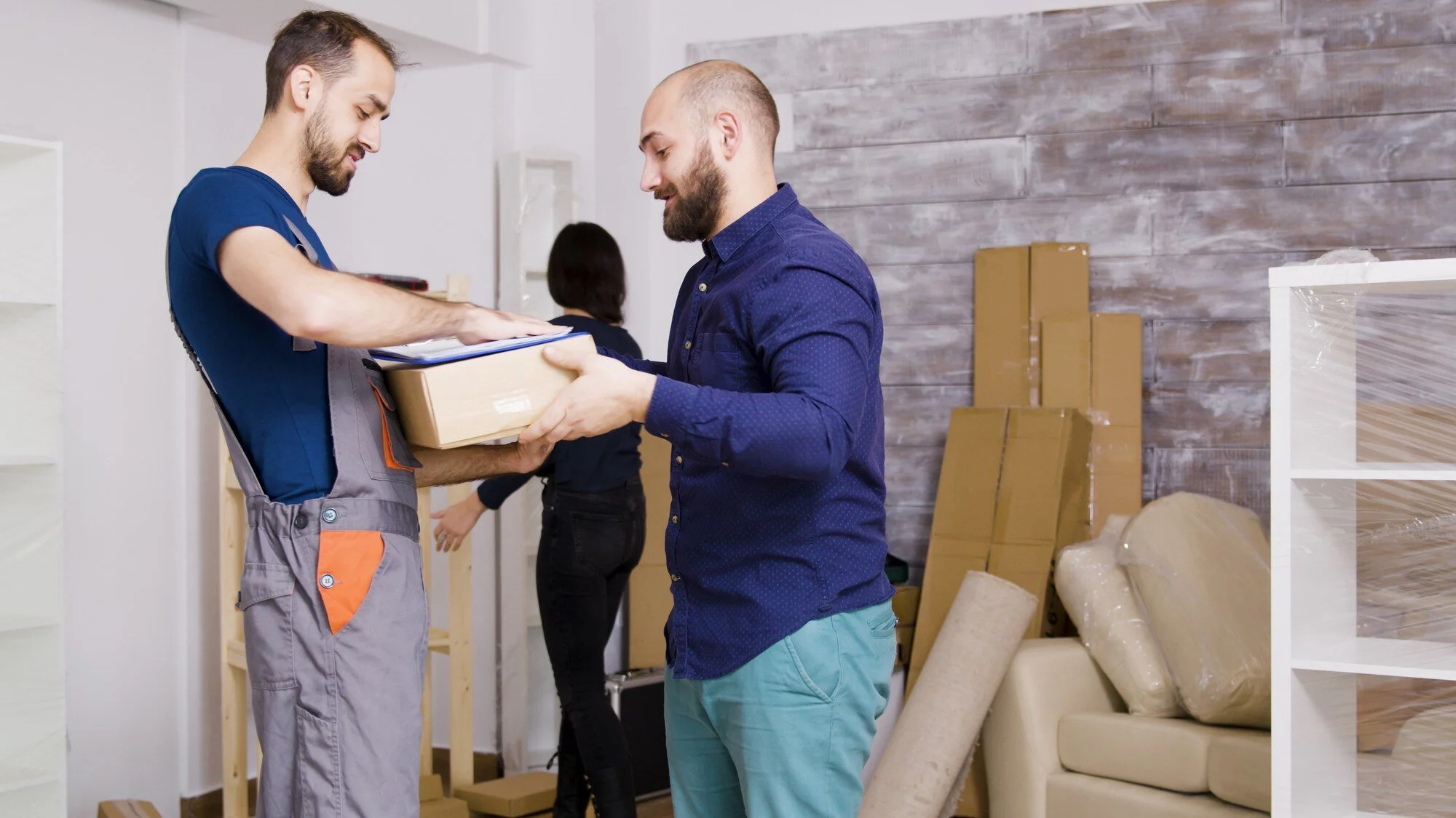 Two men exchanging a package in a room with shelves, cardboard boxes, and rolled-up packing material, indicating a warehouse or storage setting.