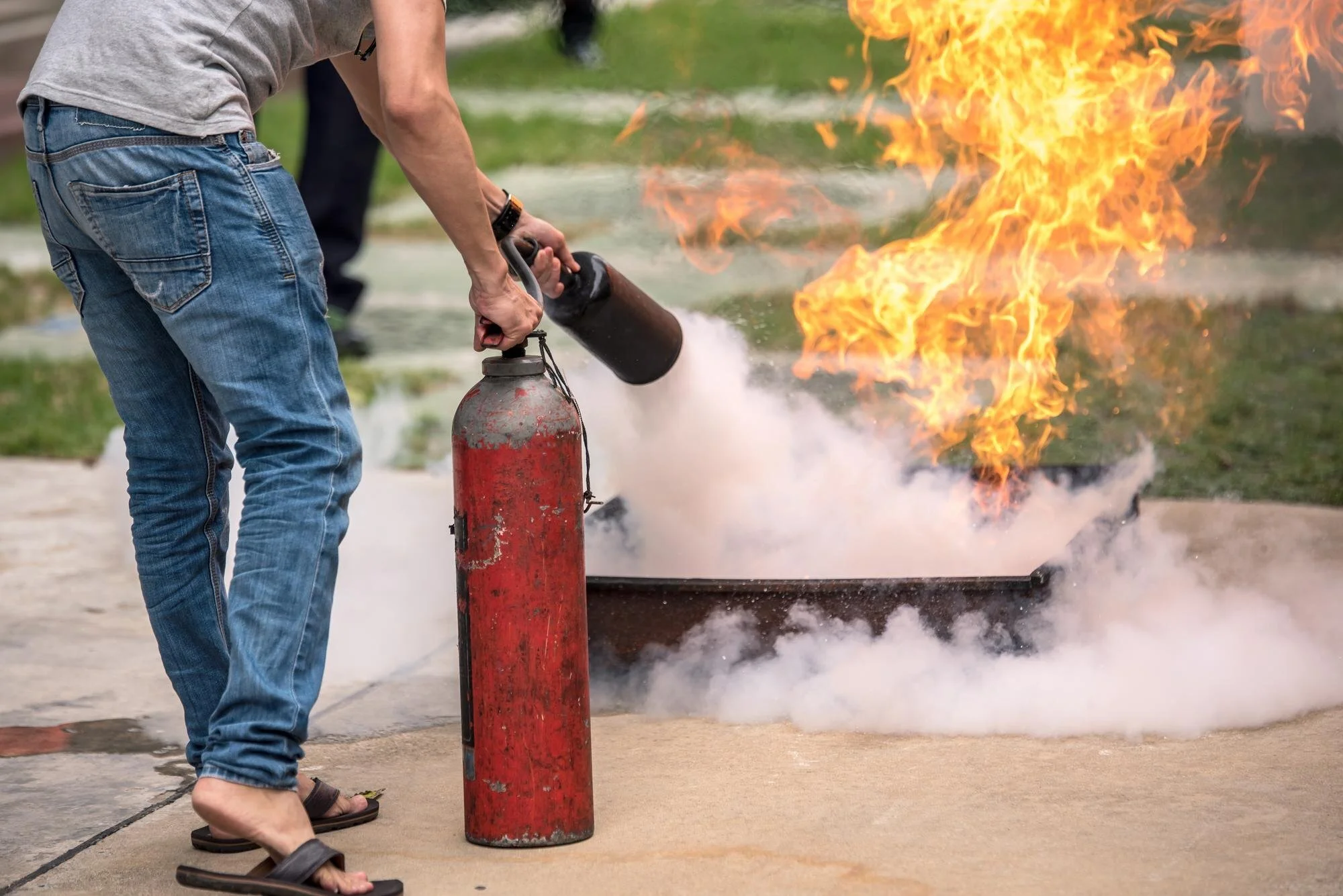 Person activating a fire extinguisher during a fire drill or emergency, with flames and smoke in the background.