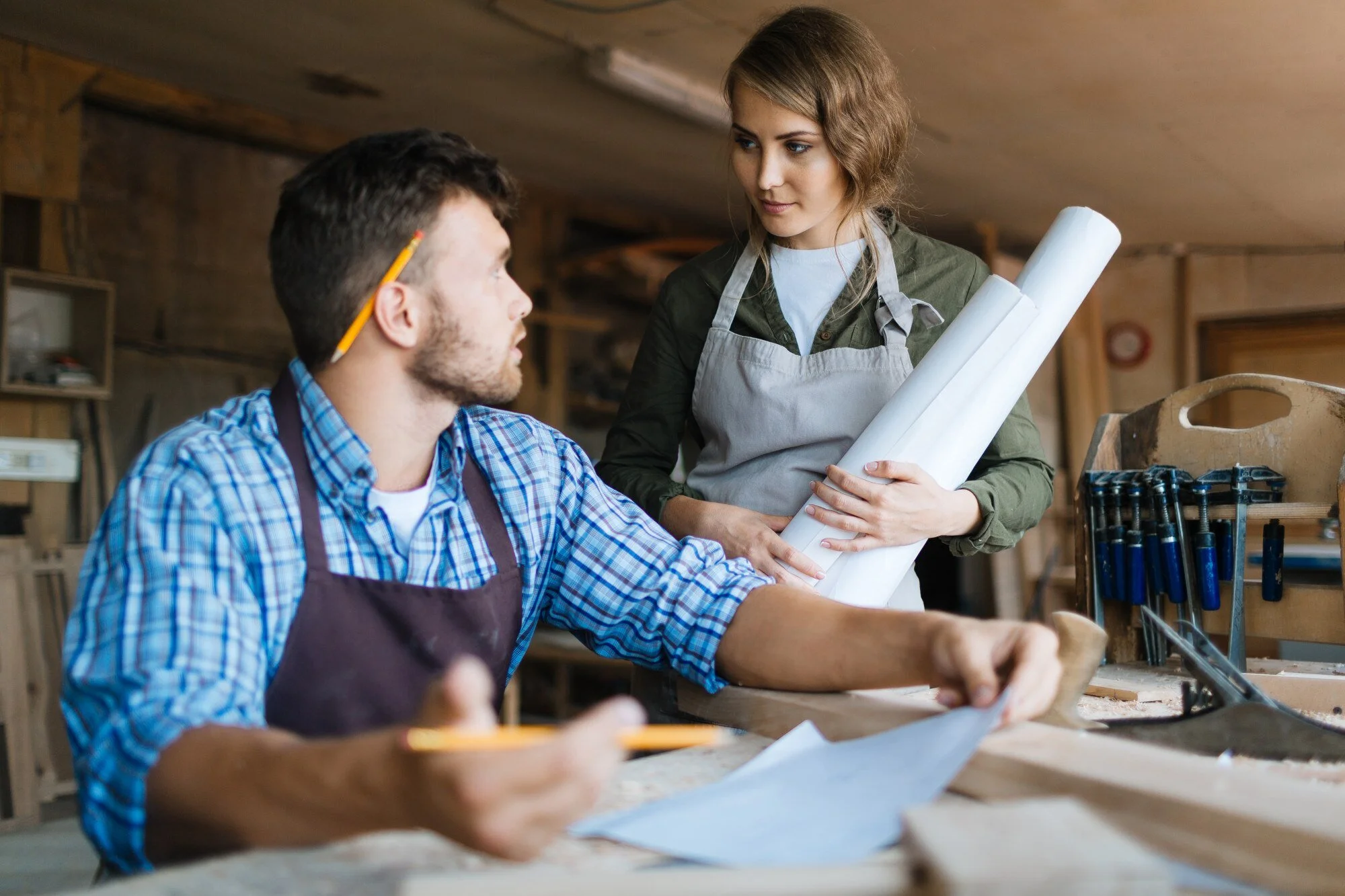 A woman holds blueprints and talks to a man in a woodworking workshop. The workshop has various tools and wood materials in the background.