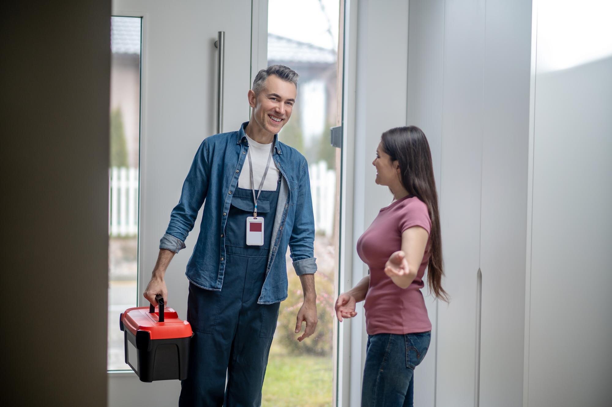 A man and a woman are having a conversation at the door of a house. The man is holding a red and black toolbox, and the woman is smiling and talking. The man wears a blue shirt and overalls with a name badge, while the woman wears a pink shirt and jeans.