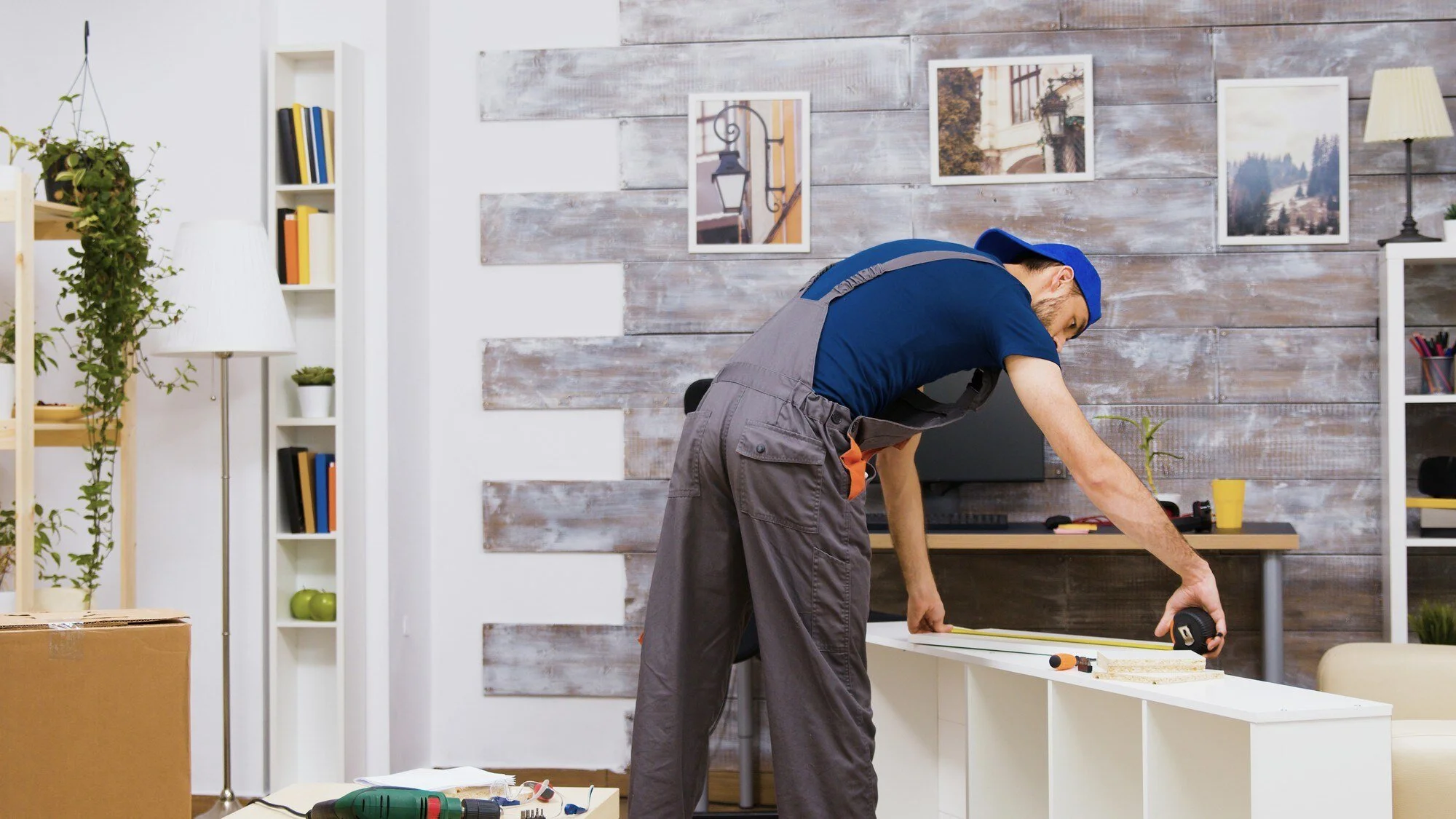 A man in work overalls measuring and working on a piece of wood at a workspace with tools, in a room with wooden wall decor and framed pictures.