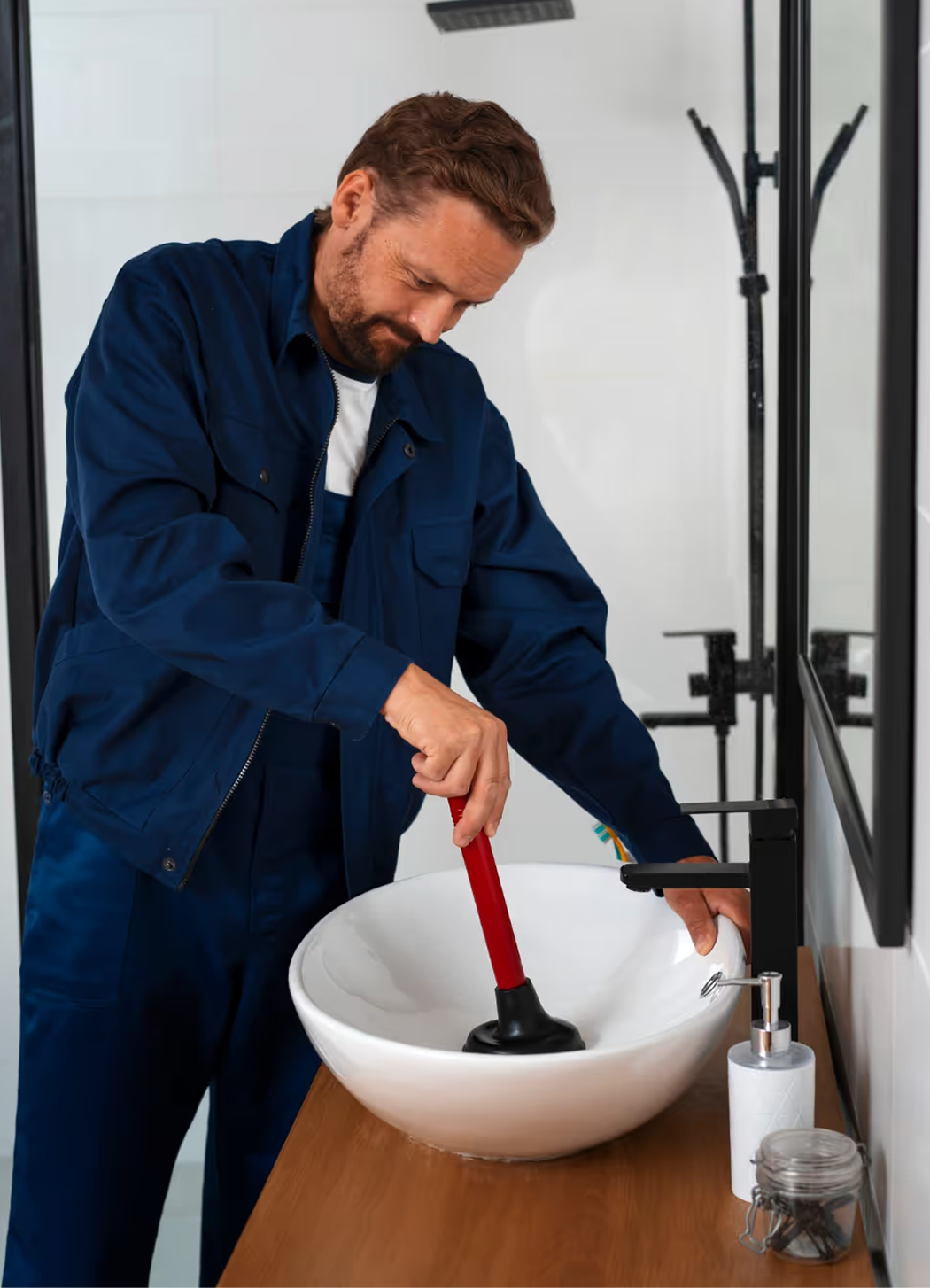 A man in a blue work uniform using a plunger to unclog a bathroom sink.