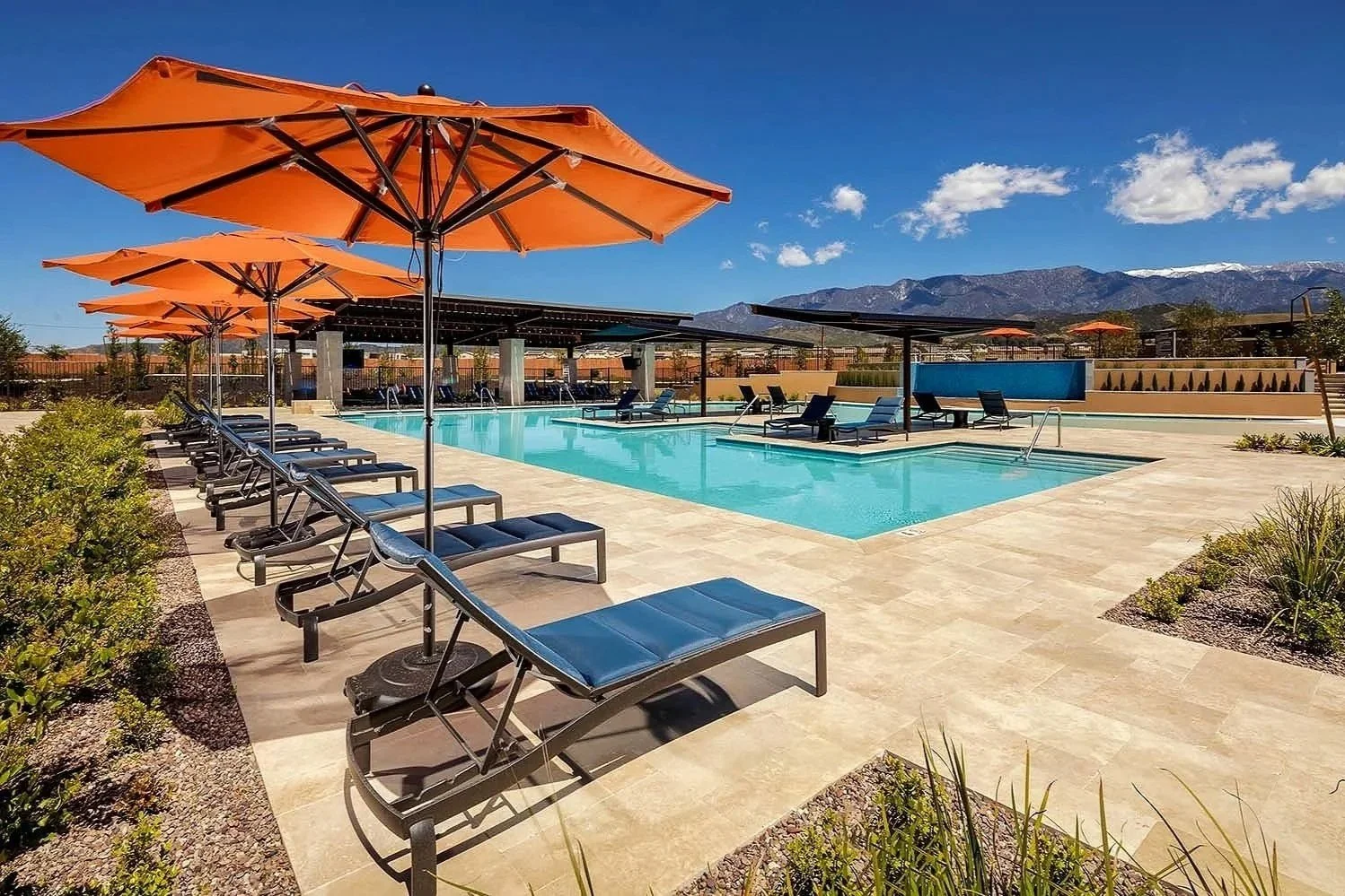 Empty outdoor swimming pool area with several black pool chairs and orange umbrellas, surrounded by desert plants and mountains in the background on a sunny day.