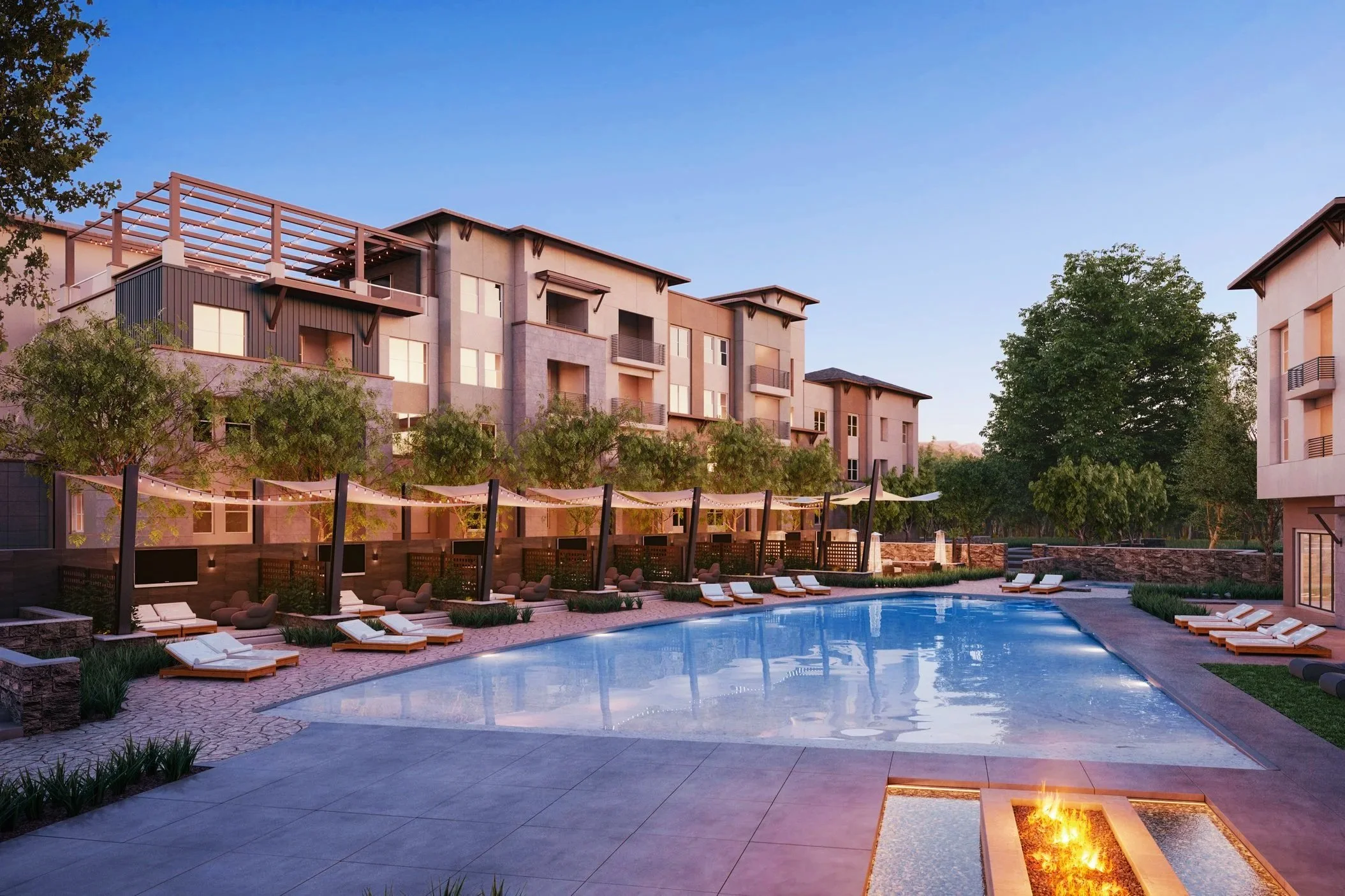 Modern apartment complex with a swimming pool, lounge chairs, and surrounding trees at sunset.