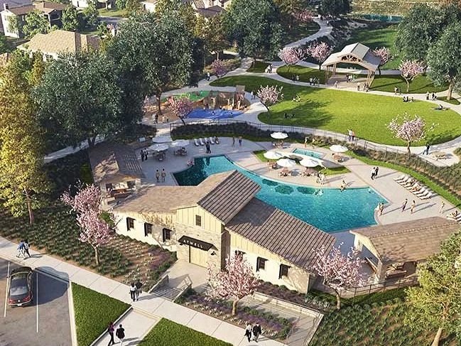 Aerial view of a community park with a swimming pool, playground, walking paths, cherry blossom trees, a grassy area, and pavilion structures, with people enjoying the outdoor space.