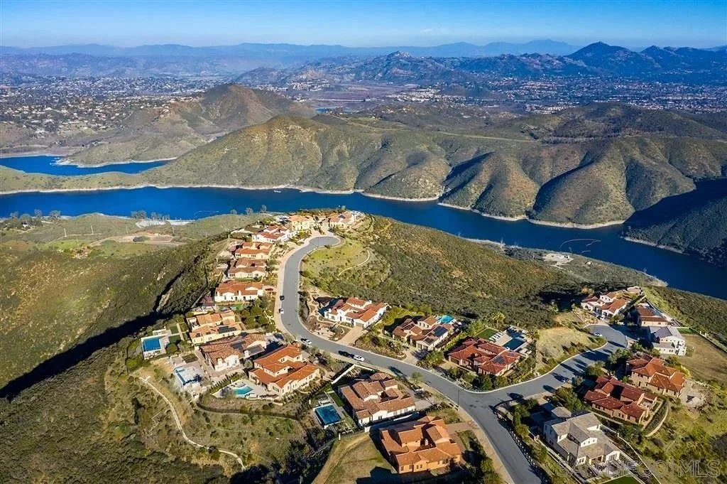 Aerial view of a residential neighborhood with houses and swimming pools on a hilly landscape, with a large blue lake and mountains in the background.