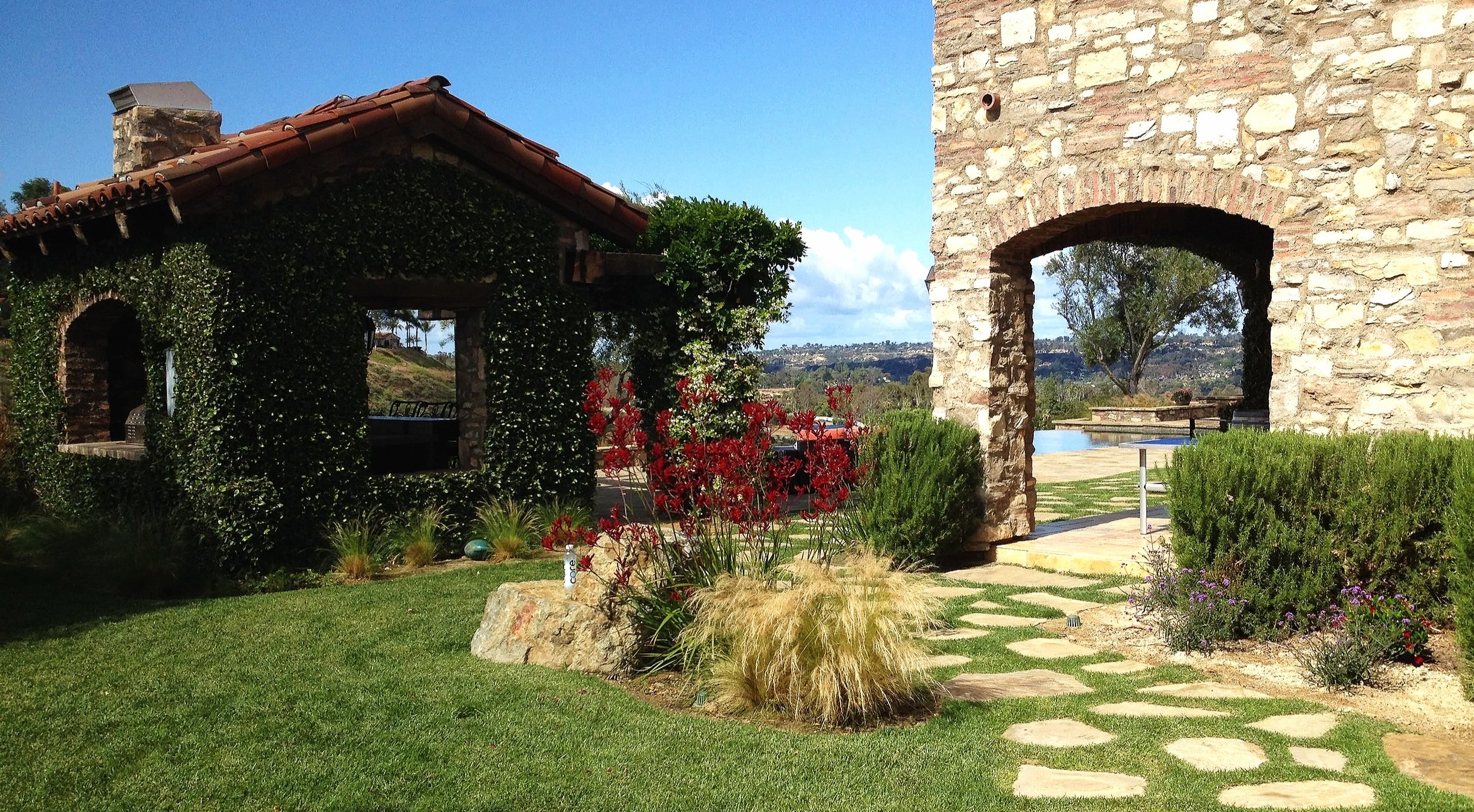 A stone archway and a small vine-covered building with a terracotta tile roof, surrounded by green grass, various plants, and flowers, with a view of a landscape and a blue sky in the background.