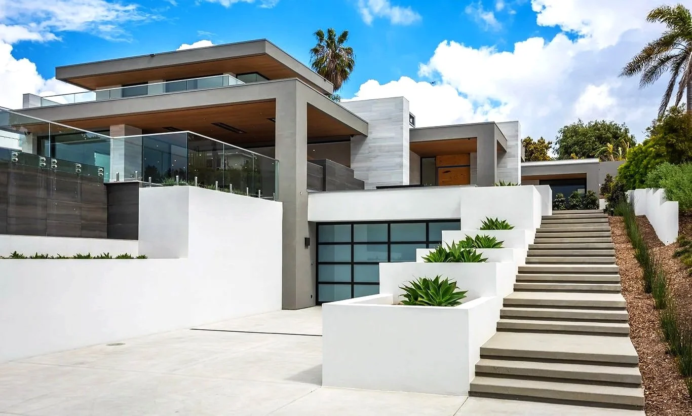 Modern multi-story house with white and gray exterior, glass balcony railings, and outdoor staircase with planters, set against blue sky with clouds and palm trees.