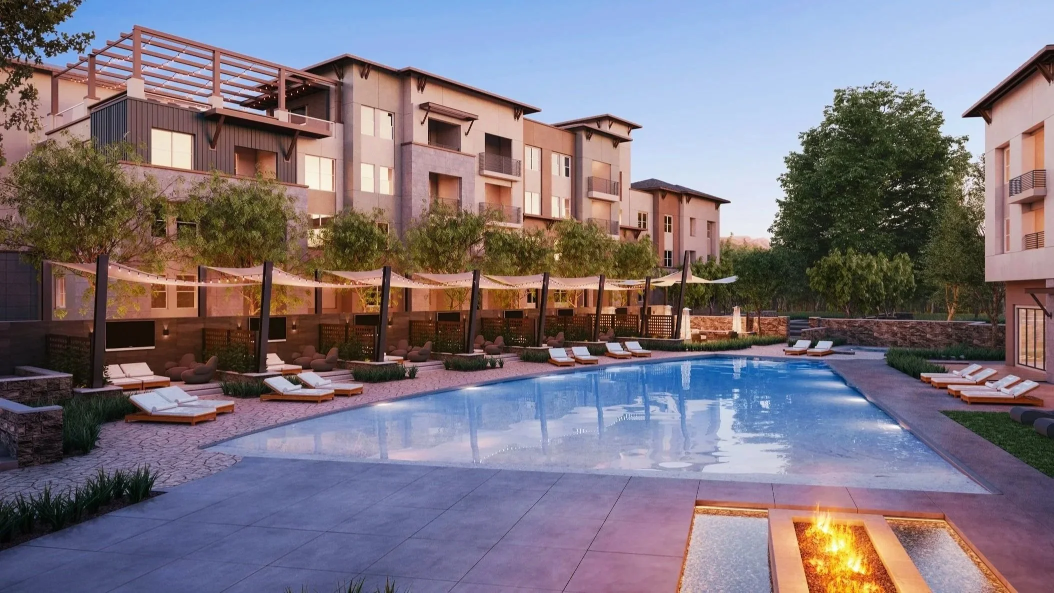 Modern apartment complex with a swimming pool, lounge chairs, and surrounding trees at sunset.