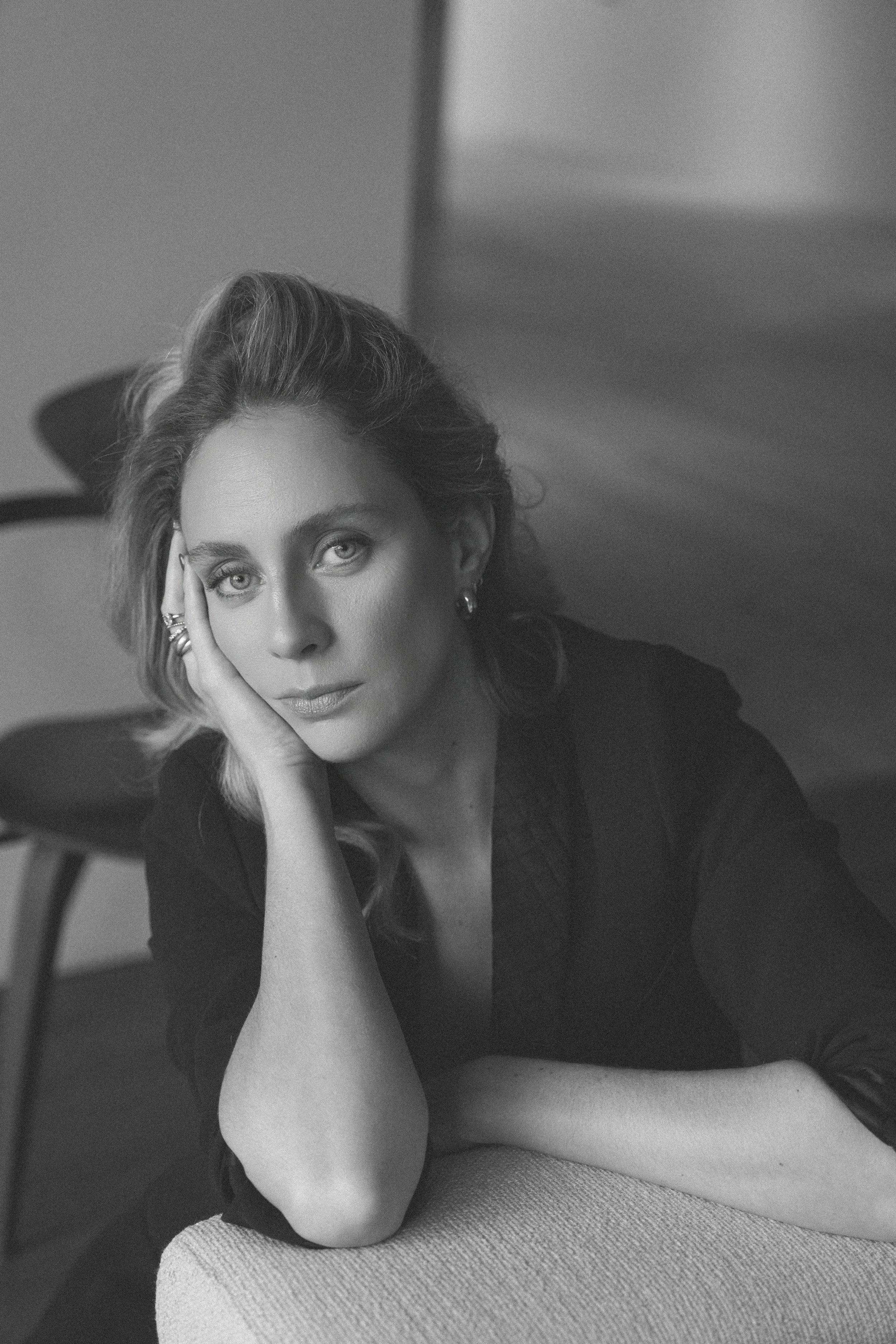 Black and white portrait of a woman with wavy hair, resting her head on her hand, looking at the camera, sitting in a room with a chair in the background.