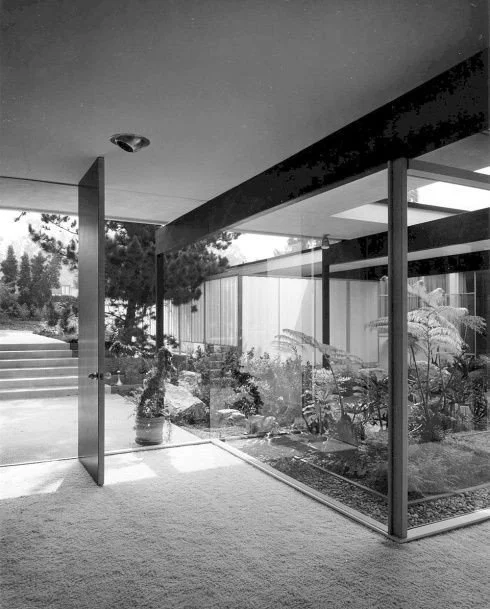 Modern house interior view with large glass windows revealing a landscaped garden outside, and a carpeted floor.