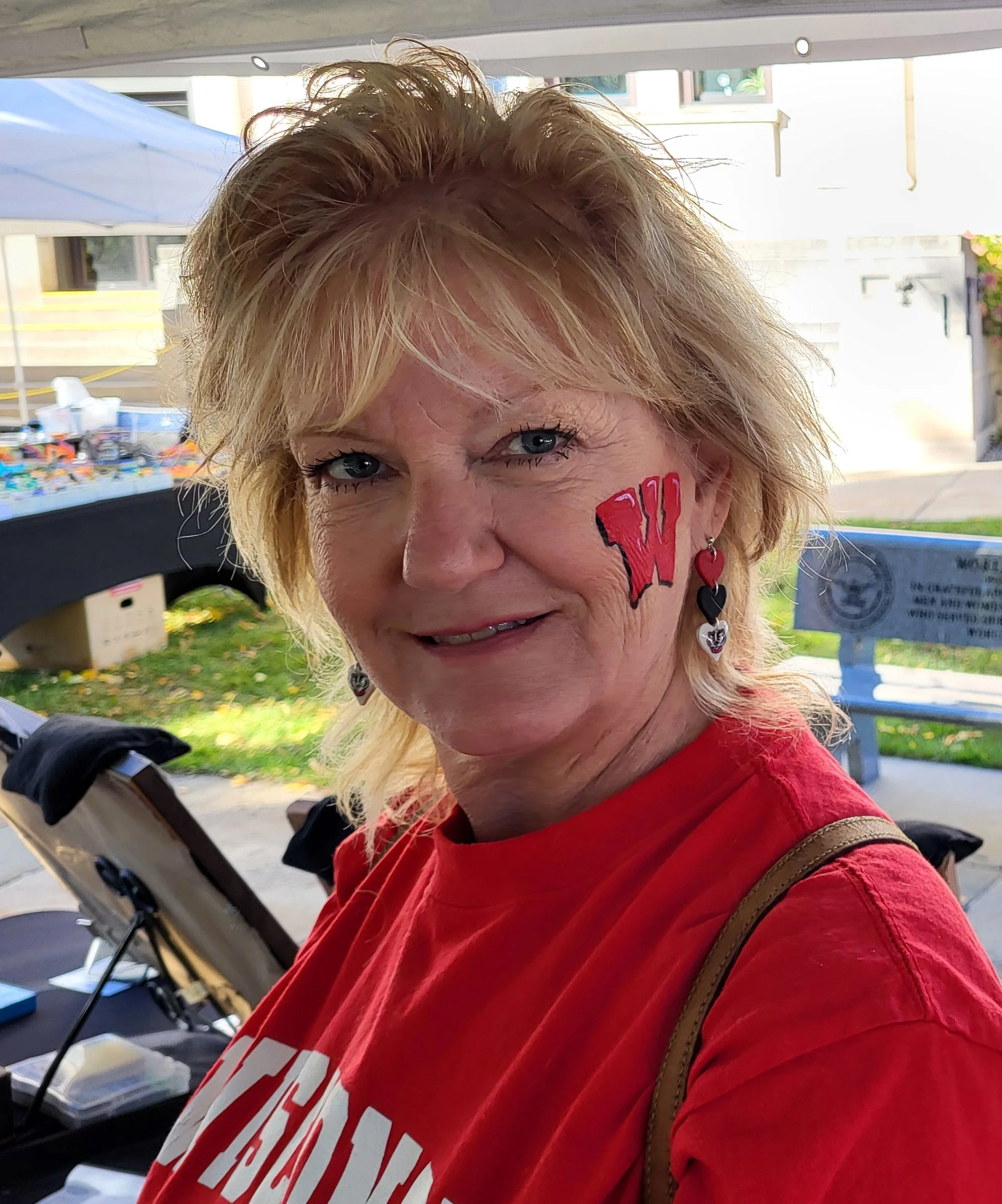 A woman in all red smiles proudly with her Wisconsin Badgers "W" face paint on her cheek that matches her earrings. The face painting was done by Kim Witte of Witty Kitty Face Painting