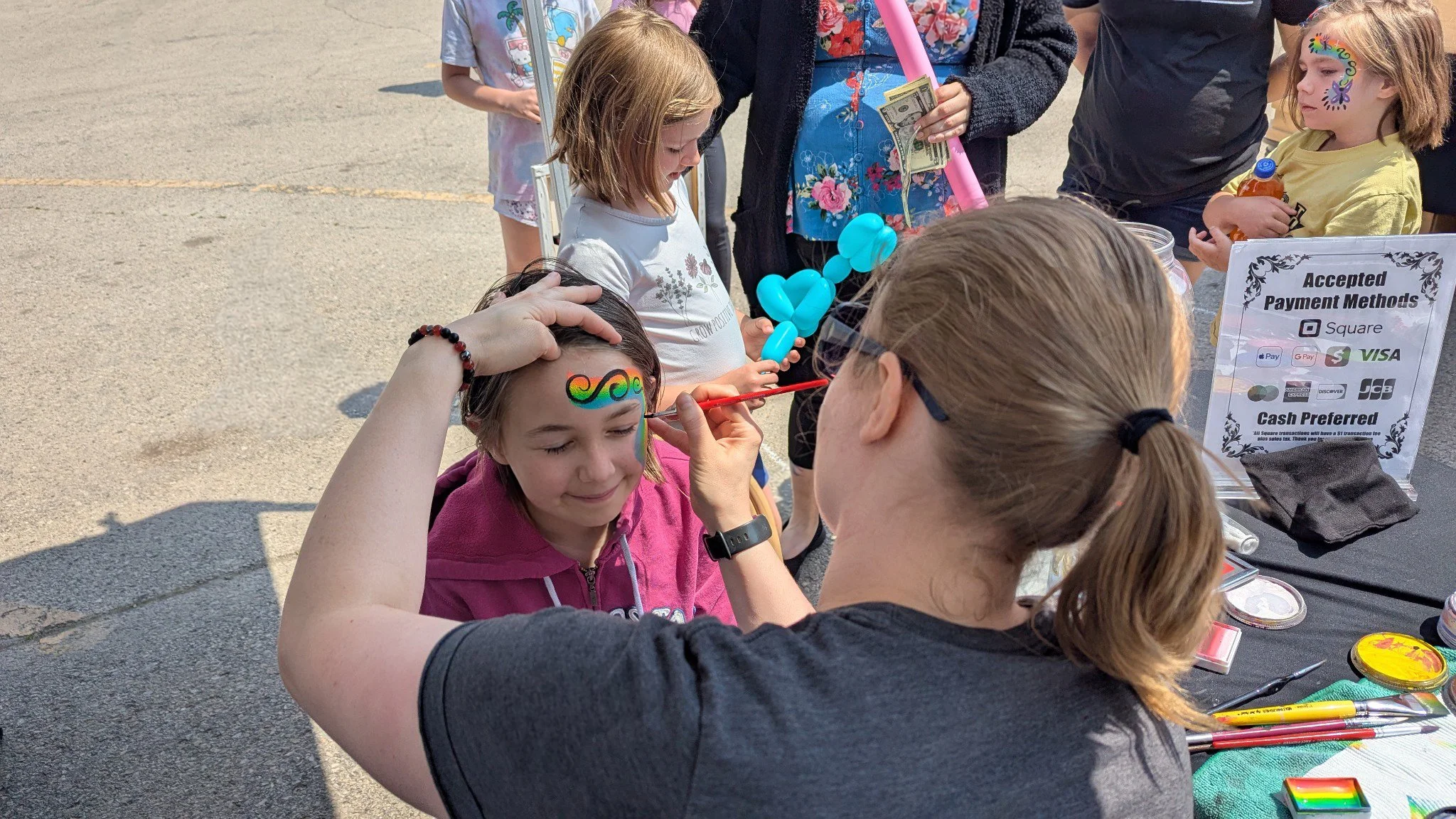 Camera looks over artist Kim's shoulder as she paints a girl with a swirly rainbow face painting. Other patrons wait in line with balloons and cash ready to get their faces painted.