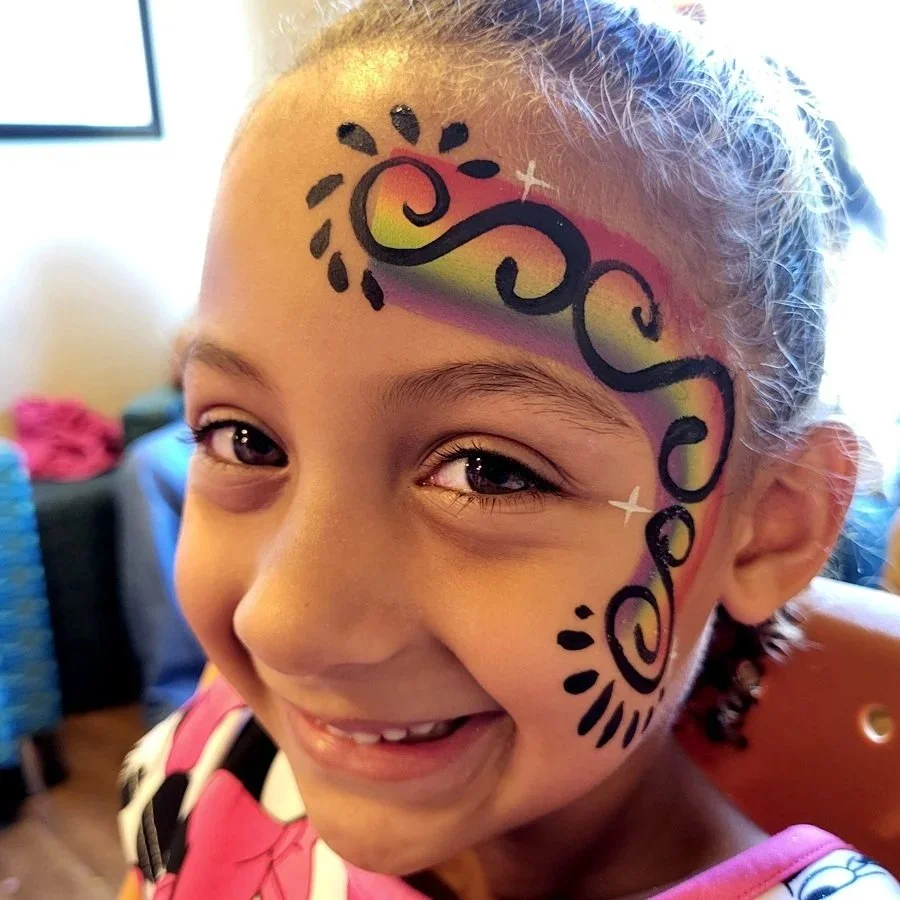 A girl at her birthday party smiles with a fun swirly sparkly rainbow face painting done by Witty Kitty Face Painting!