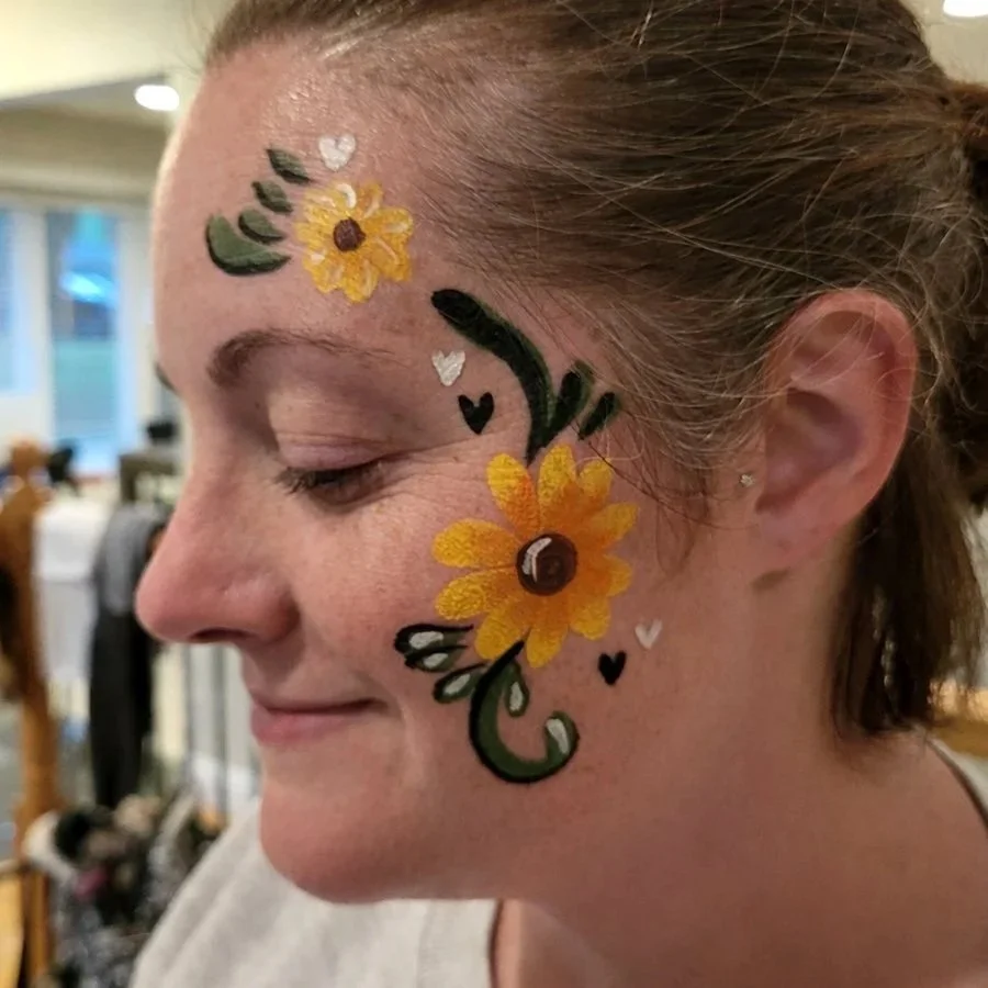 A woman smiles with a beautiful flowering face painting of sun flowers and green and black swirls.
