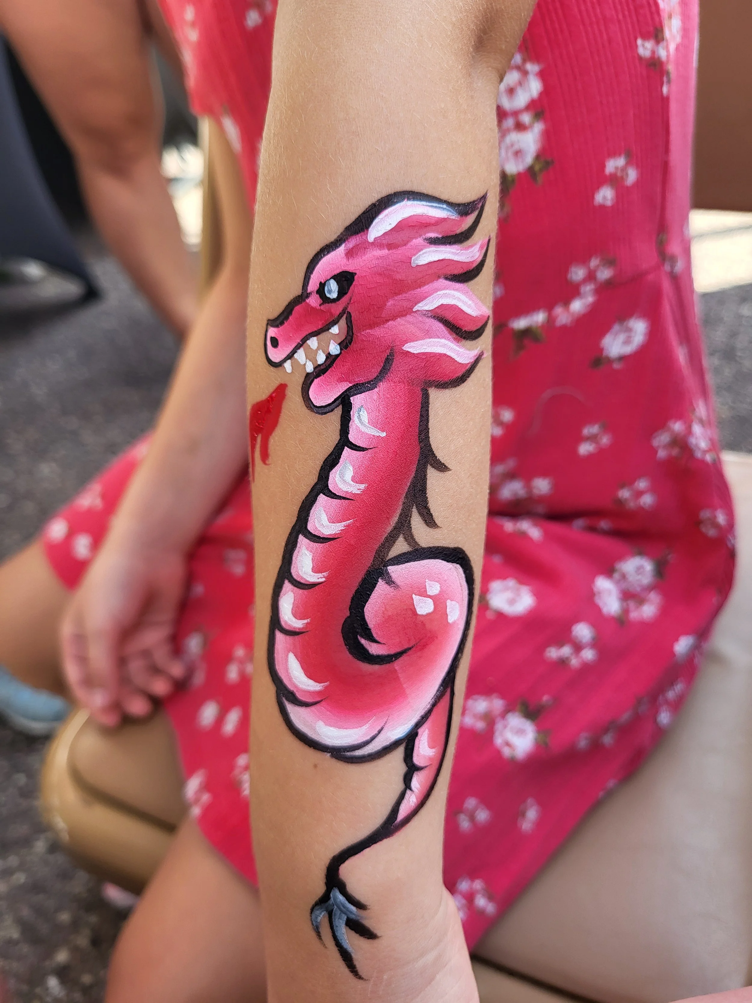 A child in a pink flowery dress sports a fancy looping dragon face paint on her arm. The dragon is white, pink, black and breathing fire. Witty Kitty Face Painting