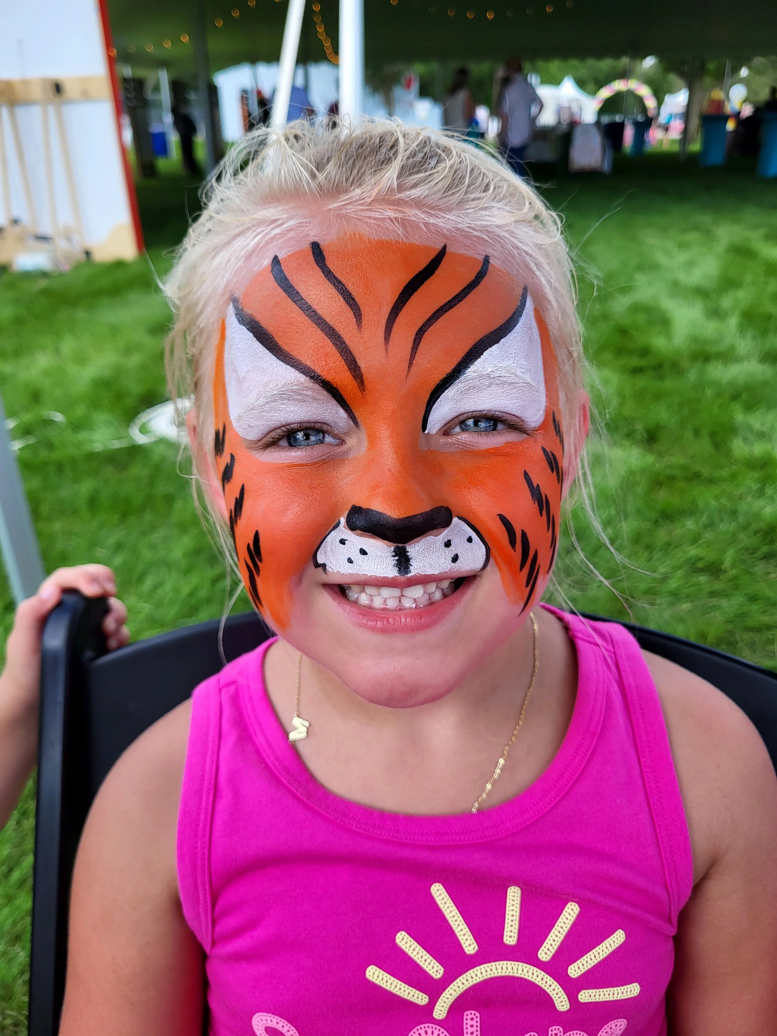 A girl smiles outside at a company picnic wearing a full face tiger face painting done by Witty Kitty Face Painting. She is wearing a pink shirt and sitting in a black chair