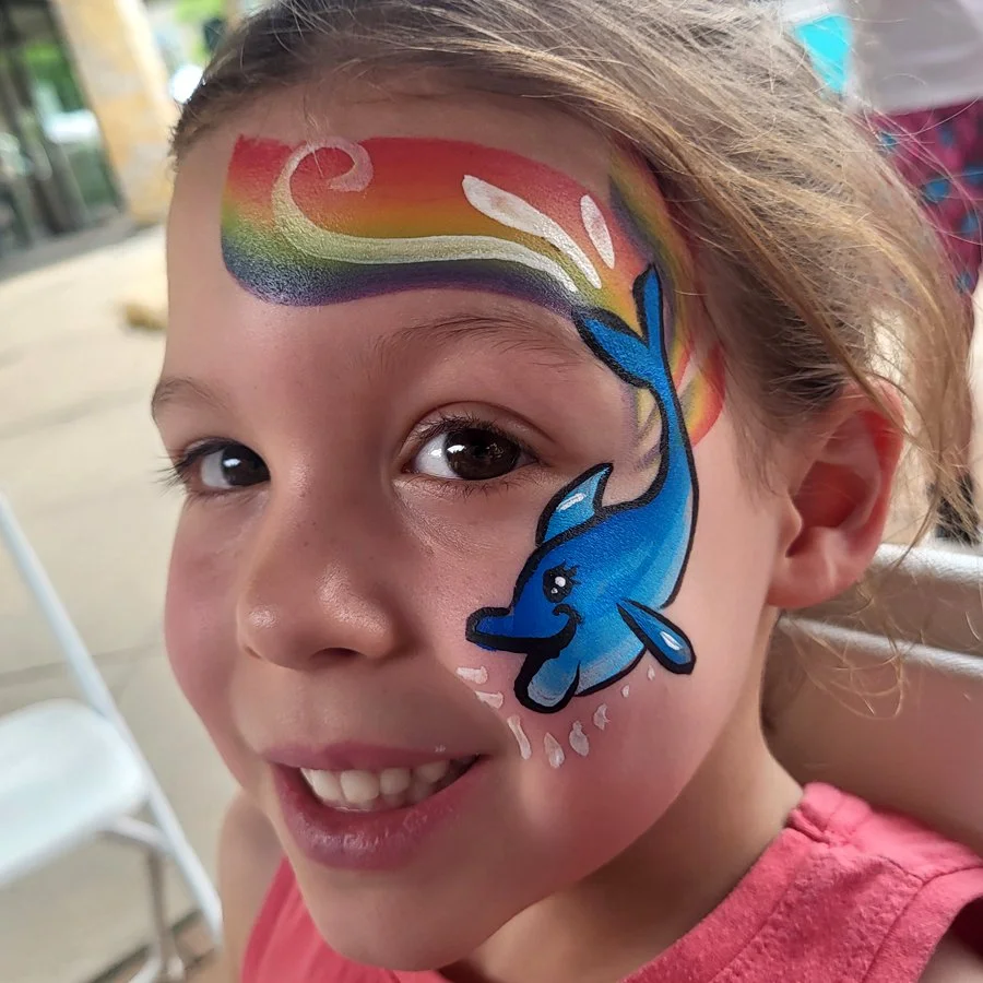 A girl smiles happily at the camera sporting her new face painting from Witty Kitty Face Painting. She has a whimsical lady dolphin who is jumping through rainbows and splashing up water. Madison Wisconsin face painting