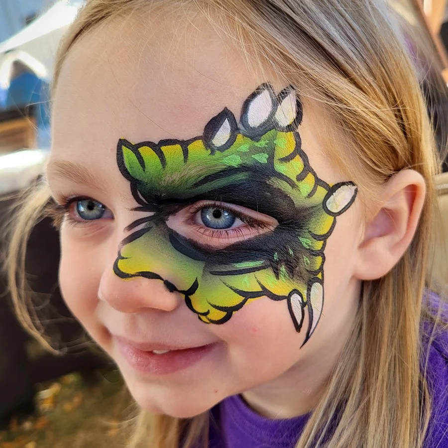 A little girl smiles excitedly with a green and yellow face painting that circles her eye. It looks like her eyes is an eye of a monster complete with horns and scales. Madison wisconsin facepainter