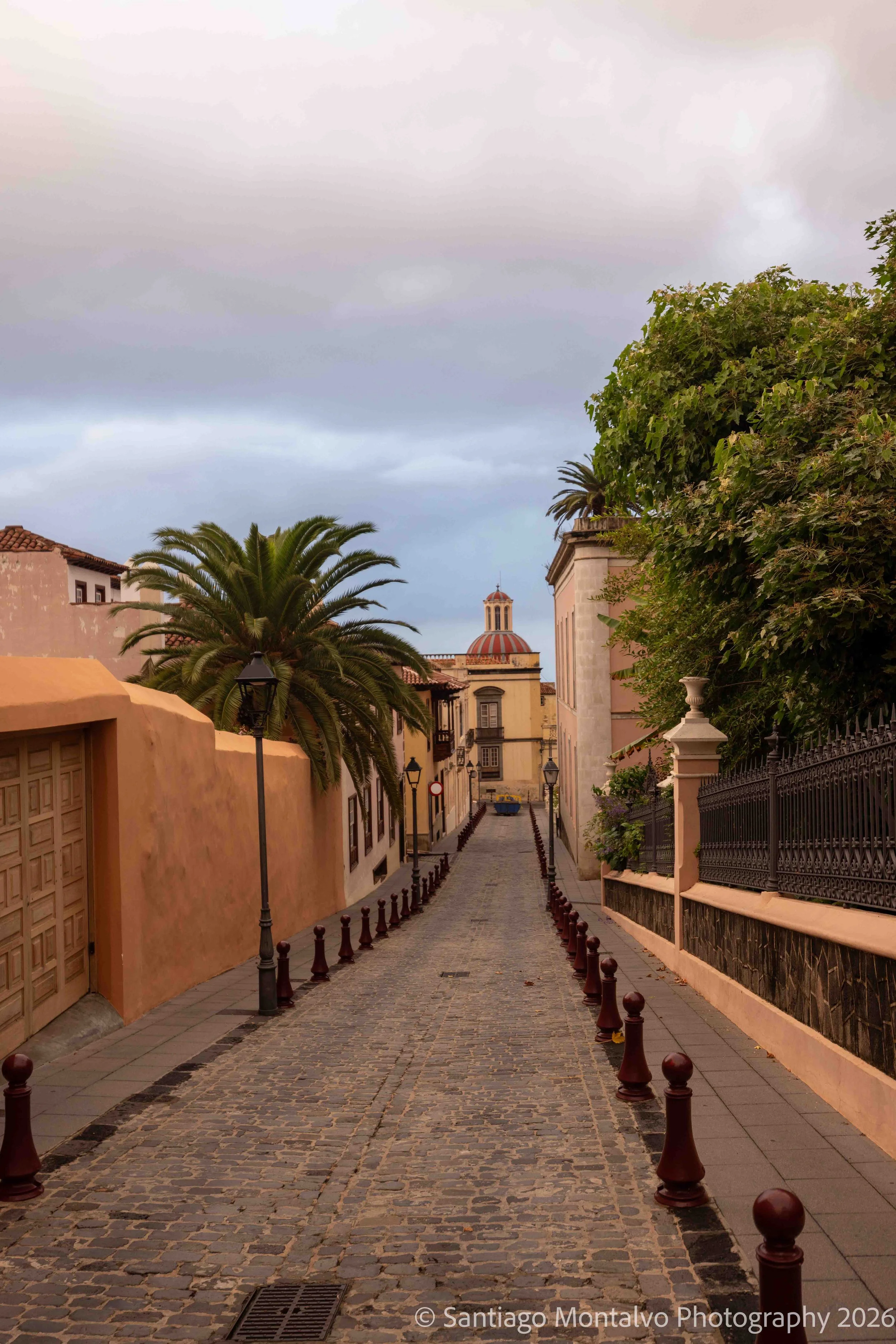 Toward the Distant Dome — Tenerife, Spain. A historic cobblestone street leads toward distinctive church architecture beneath a dramatic sky.
