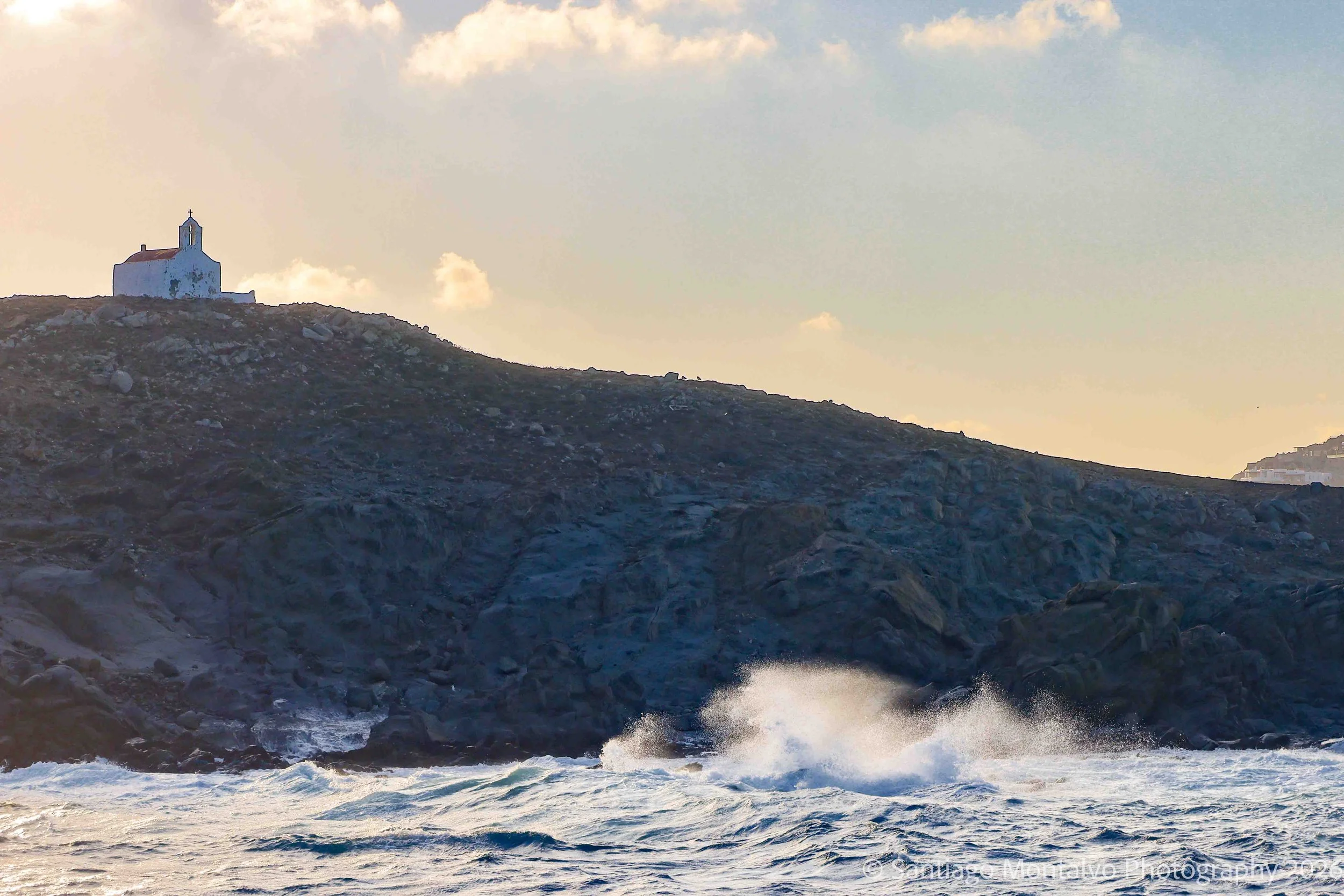 Solitude by the Sea — Mykonos, Greece. A small coastal chapel stands between rugged stone, open water, and fading light.