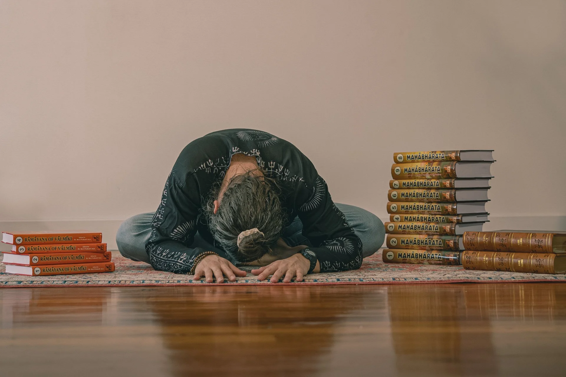 Muni Engel performing in meditation. They are surrounded by stacks of books, primarily on either side, titled 'Upanishads' 'Ramayana of Valmiki' and 'Mahabharata.'