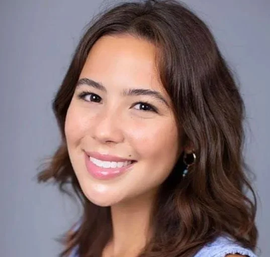 Close-up portrait of a young woman with shoulder-length brown hair, smiling, against a light blue background.