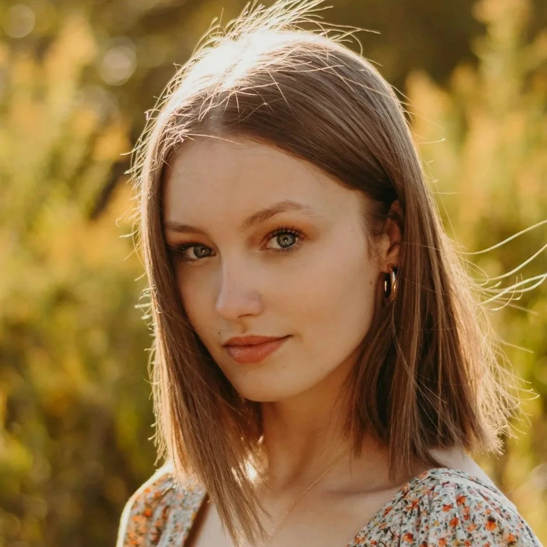 A young woman with shoulder-length brown hair and blue eyes standing outdoors in autumn sunlight.
