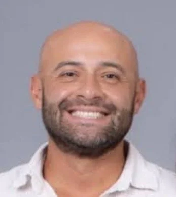 A smiling man with a shaved head and beard, wearing a white collared shirt, against a gray background.
