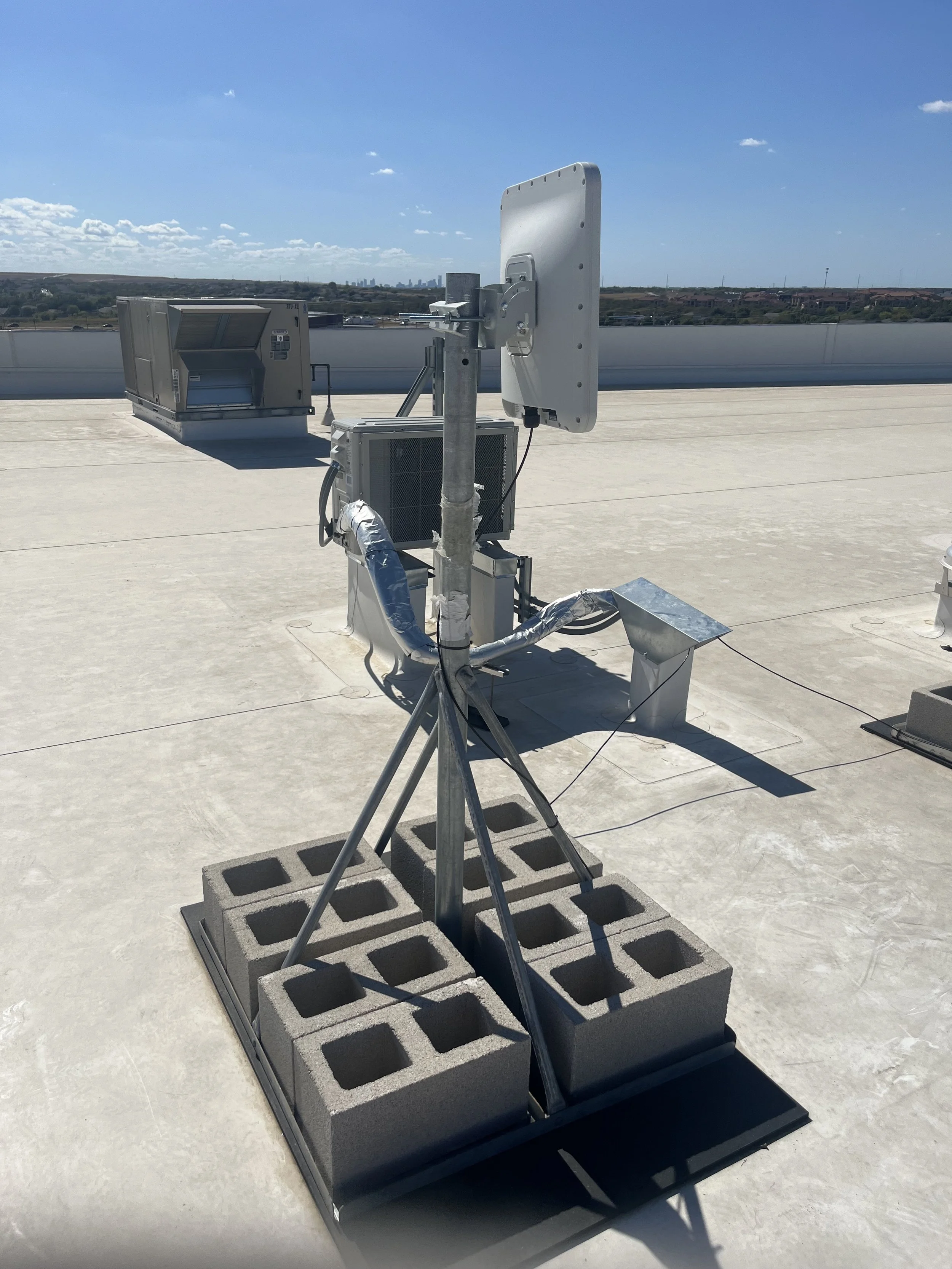 Satellite or communication equipment mounted on a rooftop with cinder blocks for stability, against a blue sky with a few clouds.