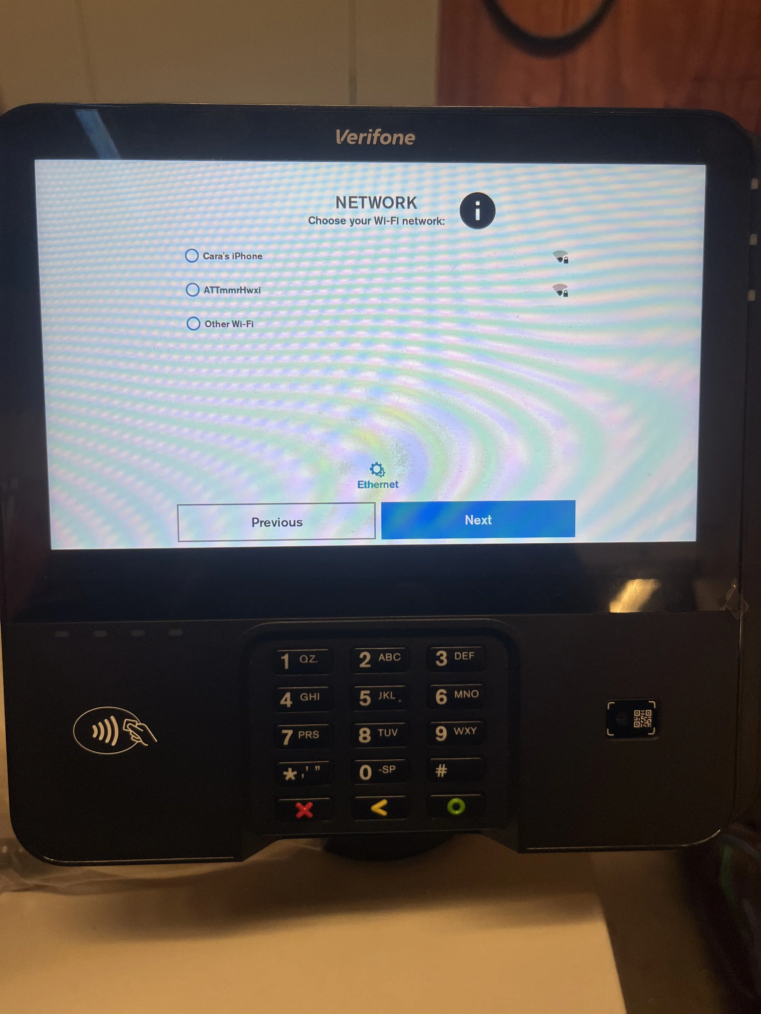 A Verifone payment terminal with a digital screen displaying Wi-Fi network options and an Ethernet icon, with a keypad below.