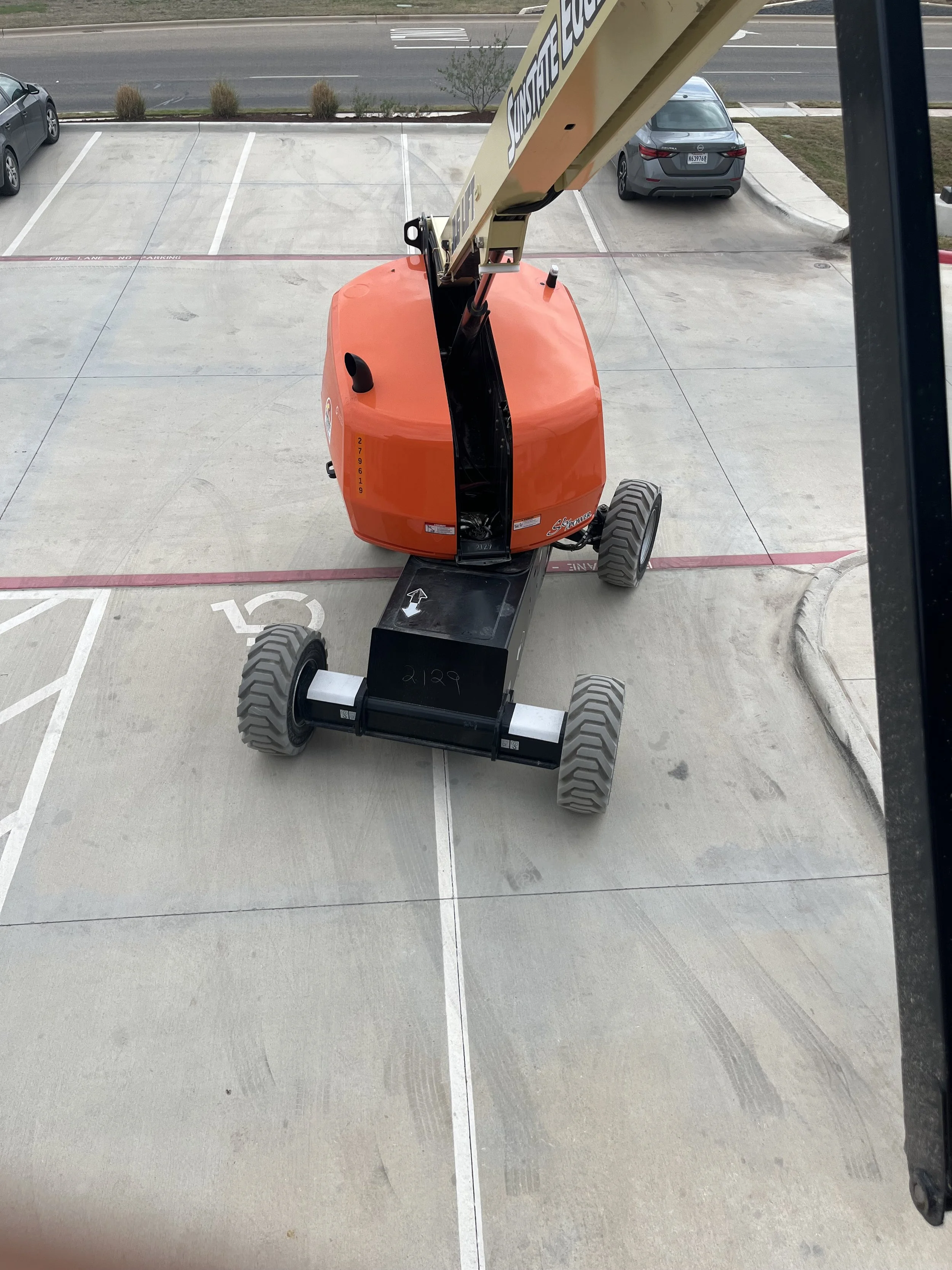 A small cherry picker lift with an orange body and black base parked in a parking lot, viewed from above.