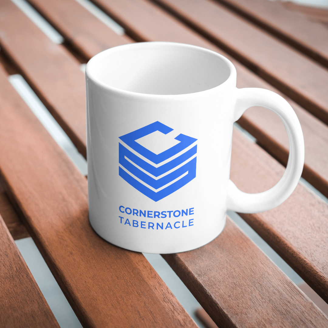 White coffee mug with blue logo and text, placed on wooden slatted surface. The logo features a cubic design and the text reads 'Cornerstone Tabernacle'.