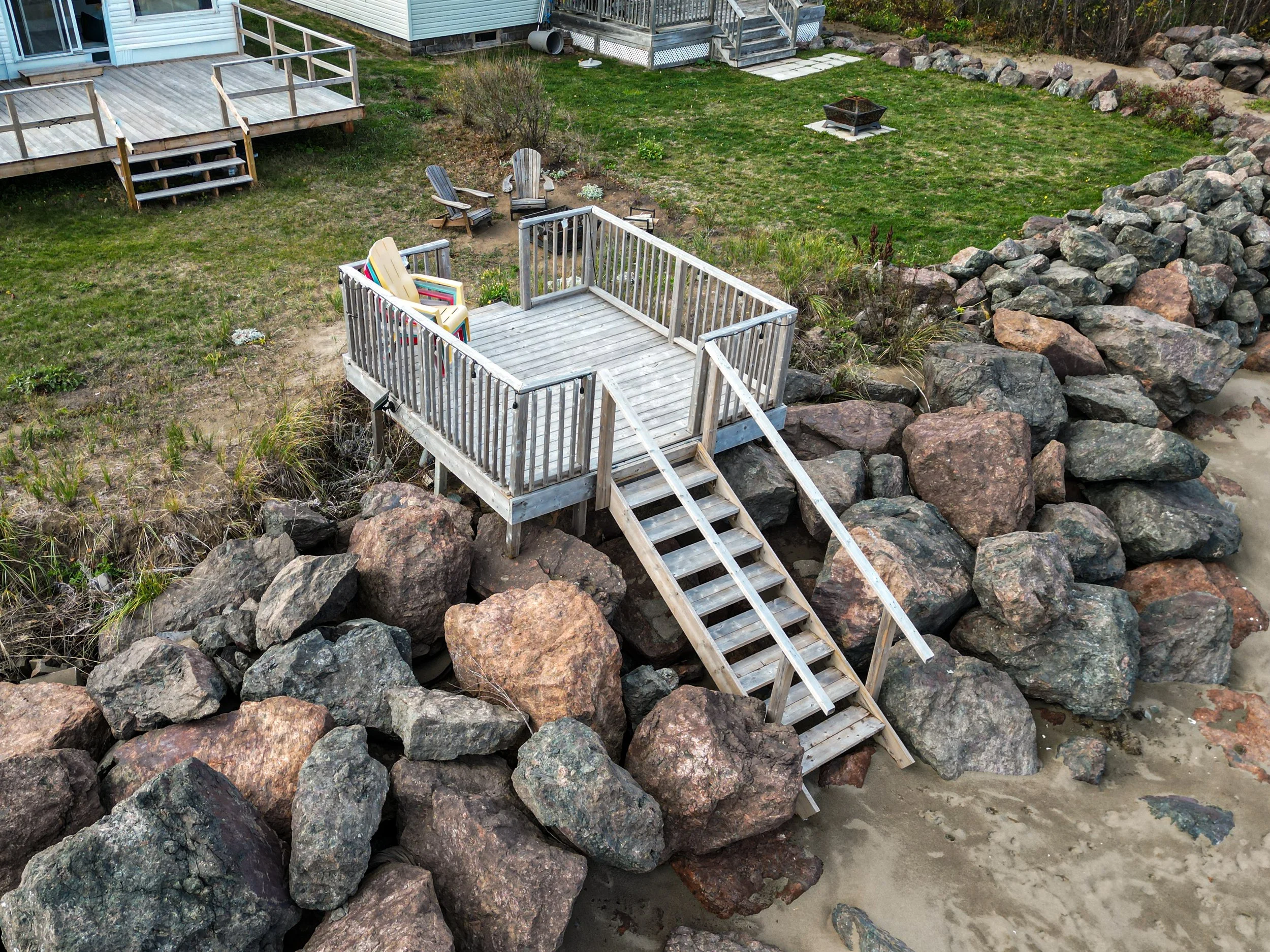 A small wooden dock with stairs leads down to the sandy beach, surrounded by large rocks. The dock has a couple of chairs and an Adirondack chair with a towel draped over it. There's a grassy backyard with a deck and some outdoor furniture in the background.