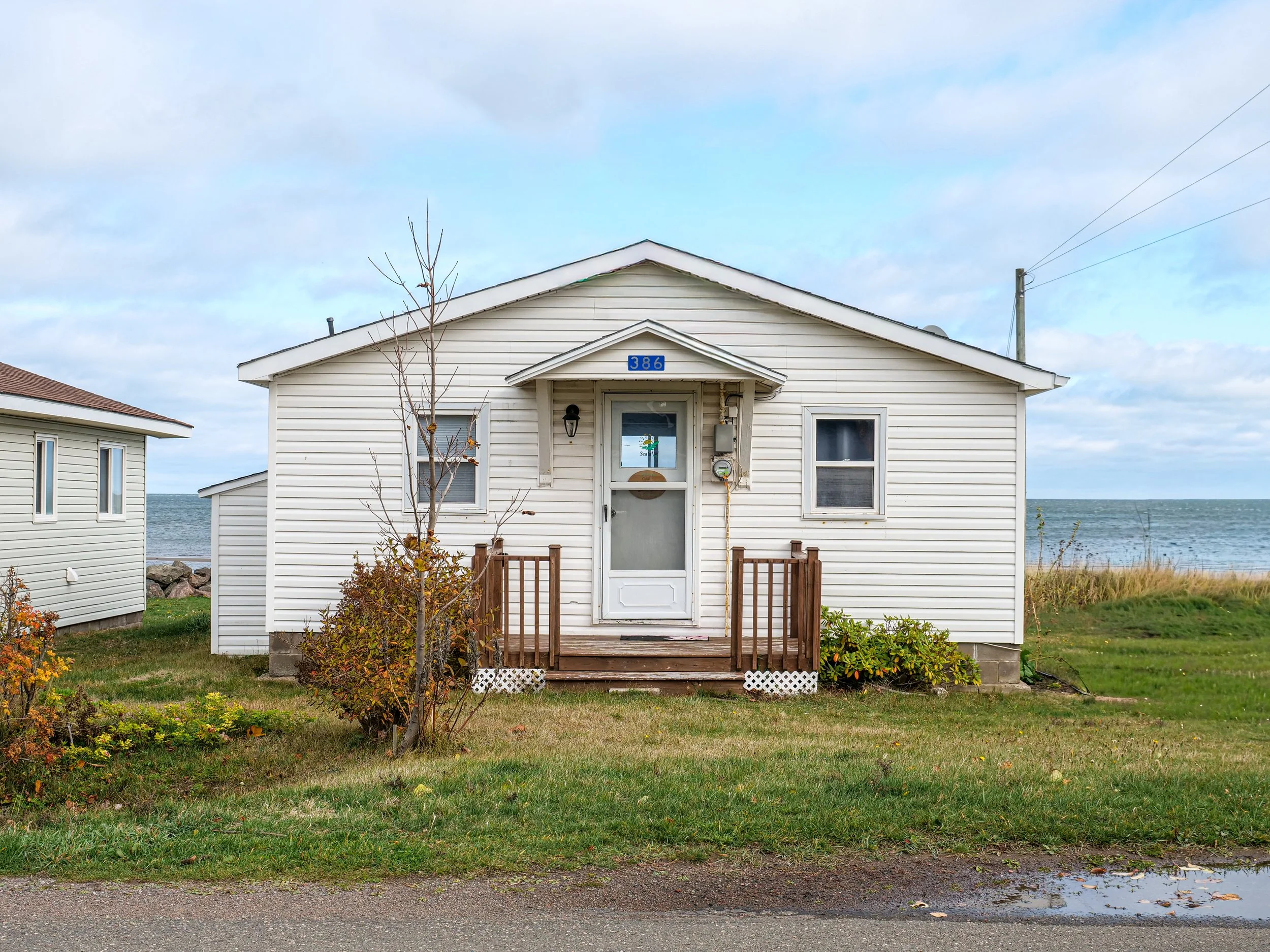 A white house with a small front porch and steps, located near the beach with ocean visible in the background, and a small tree and bush in the yard.
